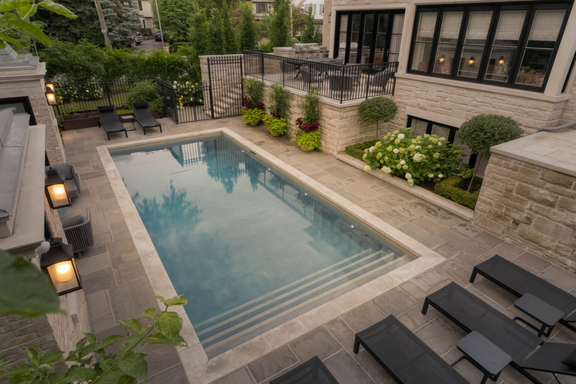 A serene backyard with a rectangular pool, surrounded by lounge chairs, greenery, and a stone-paved area adjacent to a modern house.