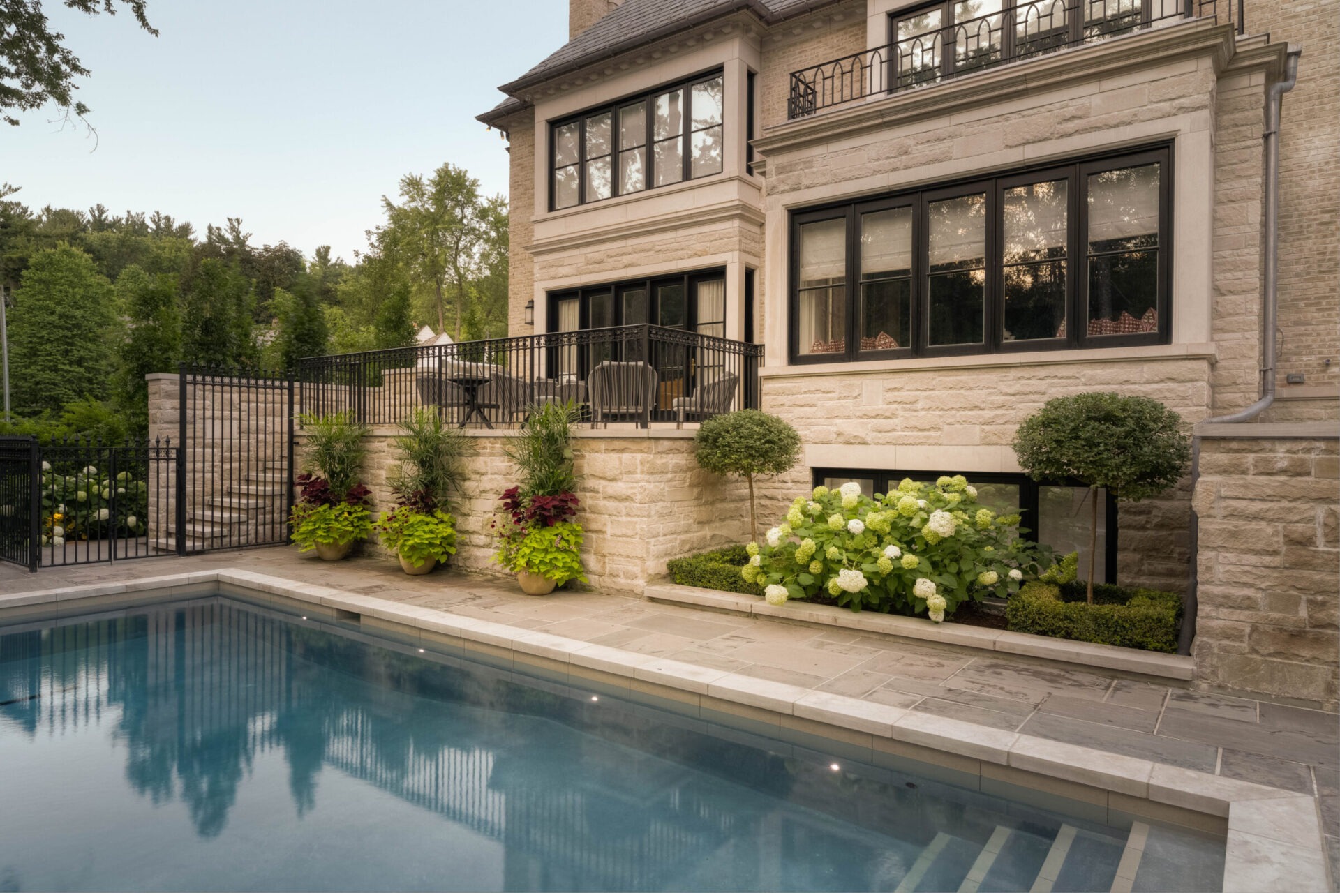 Elegant stone house with large windows overlooks a serene pool, surrounded by landscaped greenery and patio furniture, set against a lush forest backdrop.