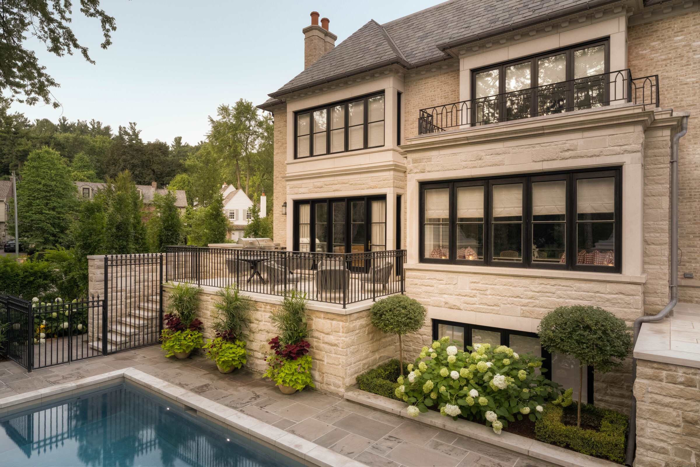 Elegant stone house with large windows, balcony, and patio overlooking a pool. Lush greenery surrounds the area, creating a serene atmosphere.