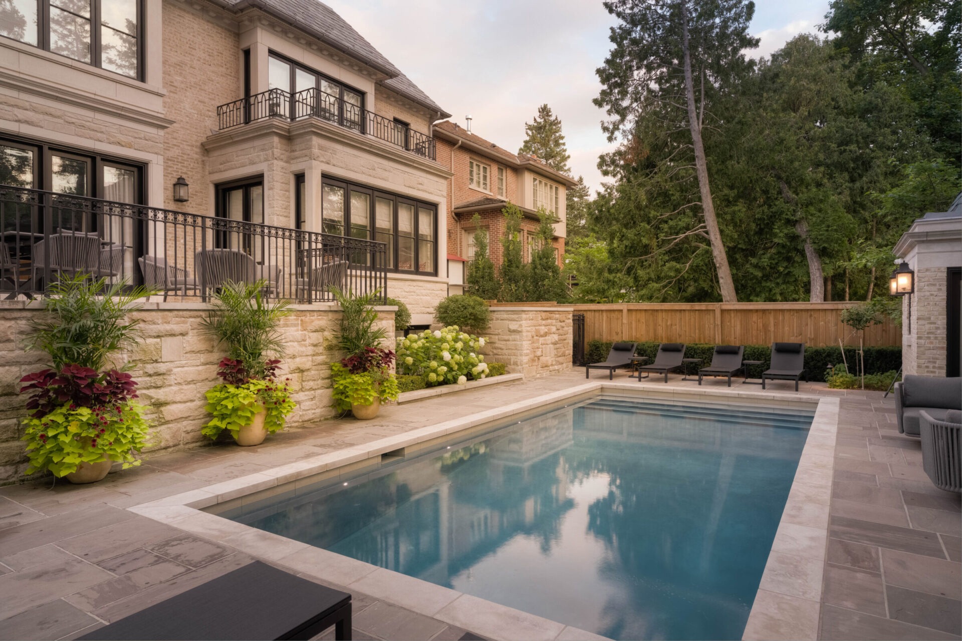 Elegant backyard with a modern stone house, pool, and lush greenery. Outdoor seating and decorative plants enhance the relaxing atmosphere.