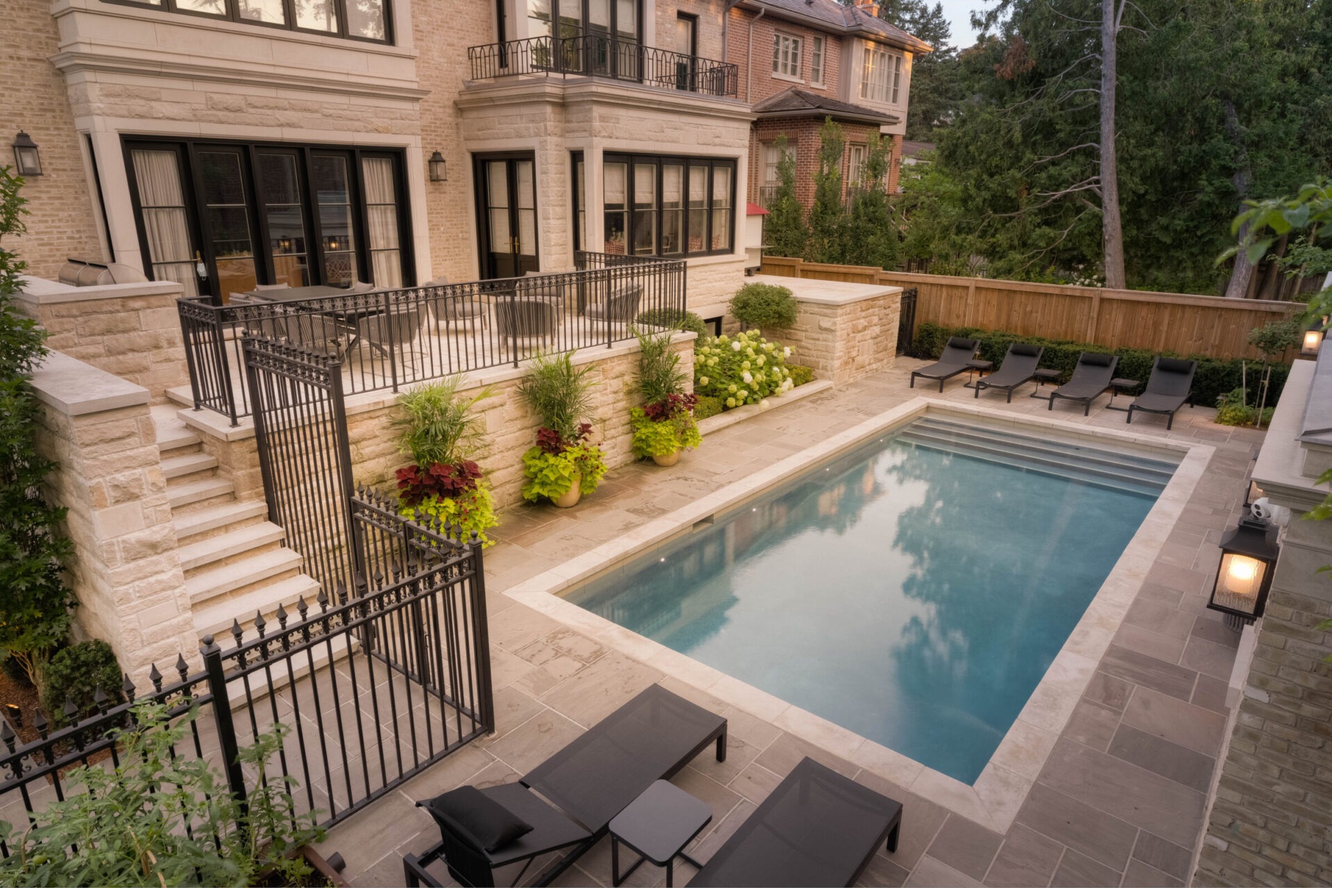 Elegant backyard features modern pool, stone patio, lush greenery, and stylish loungers. Large windows overlook the serene space, ideal for relaxation.