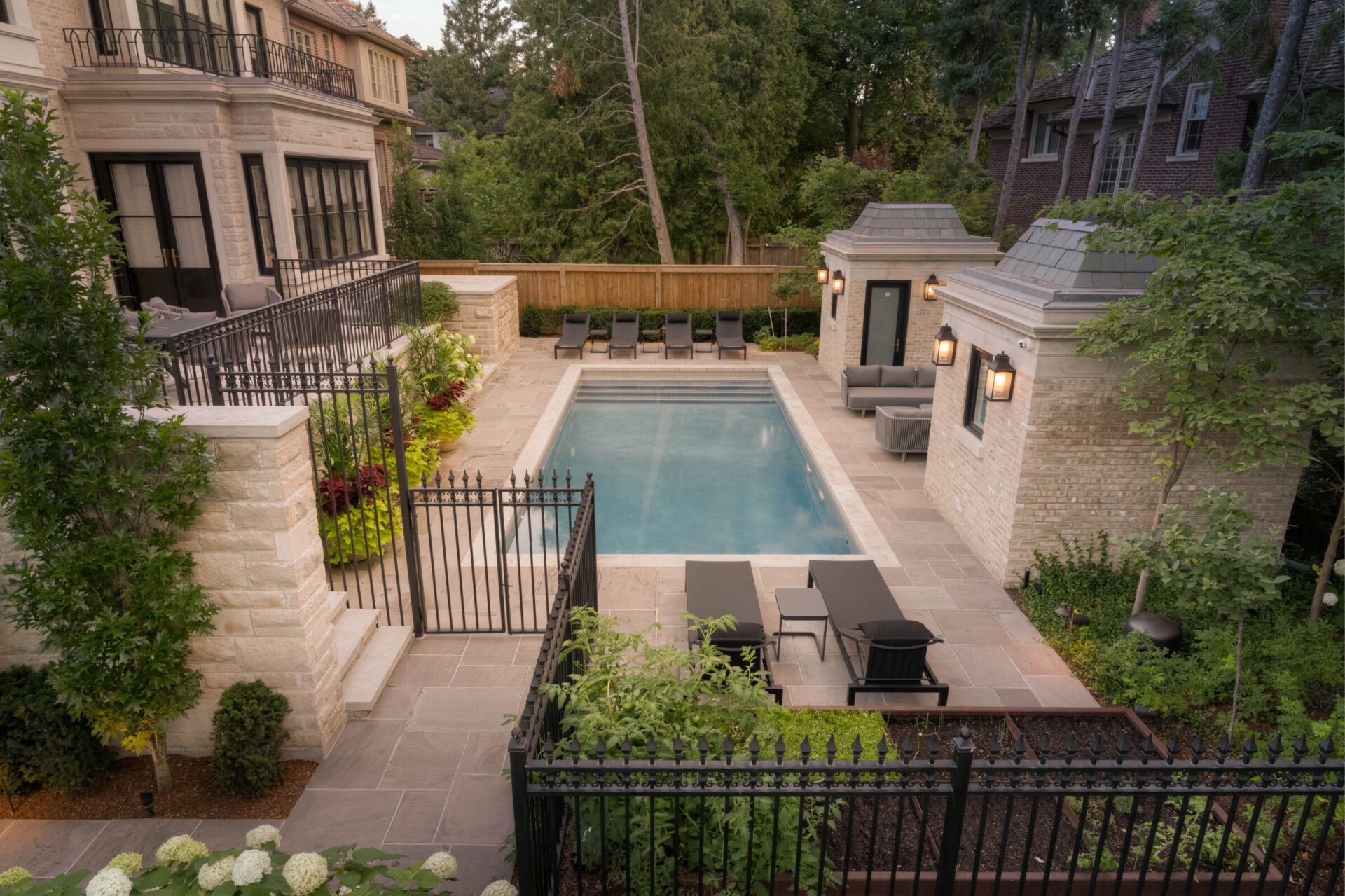 A serene backyard features a rectangular swimming pool, surrounded by greenery, lounge chairs, and elegant stone architecture, with tall trees in the background.