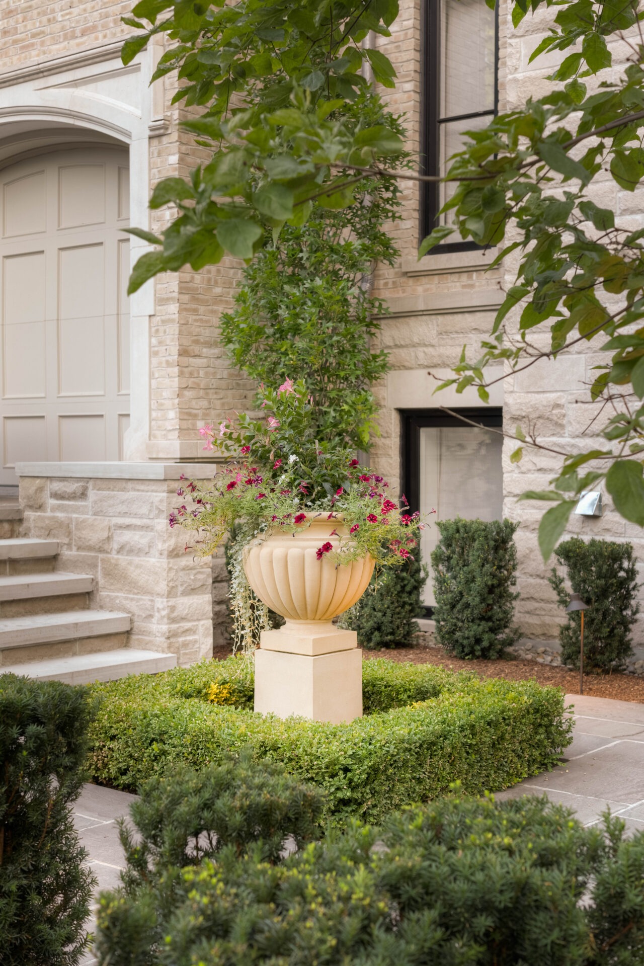 Elegant stone planter with pink flowers in a neatly manicured garden, flanked by shrubs, beside a brick home's entrance.