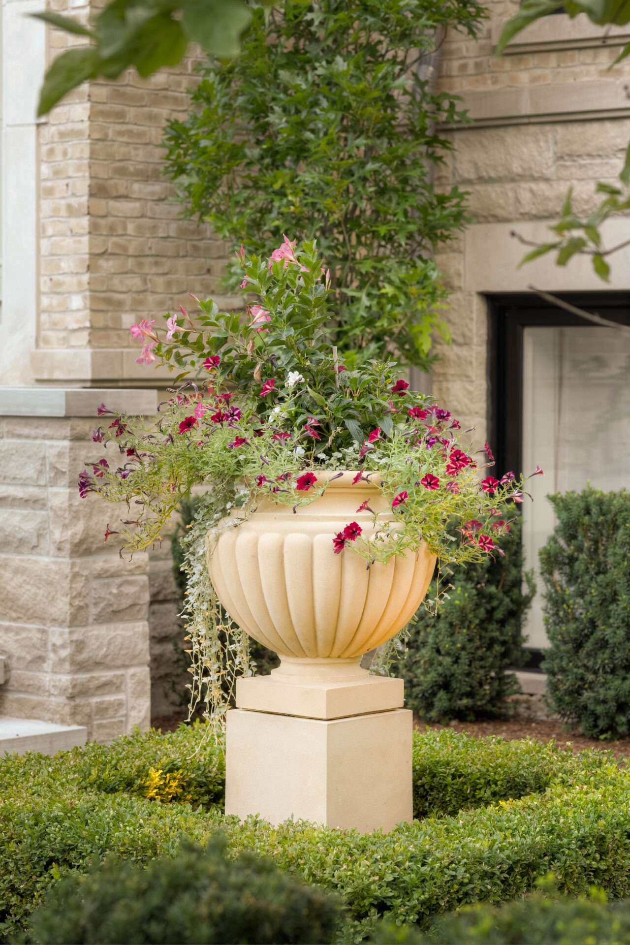 Elegant stone planter with vibrant flowers amidst manicured greenery, set against a classic brick and stone building facade. Peaceful garden scene.