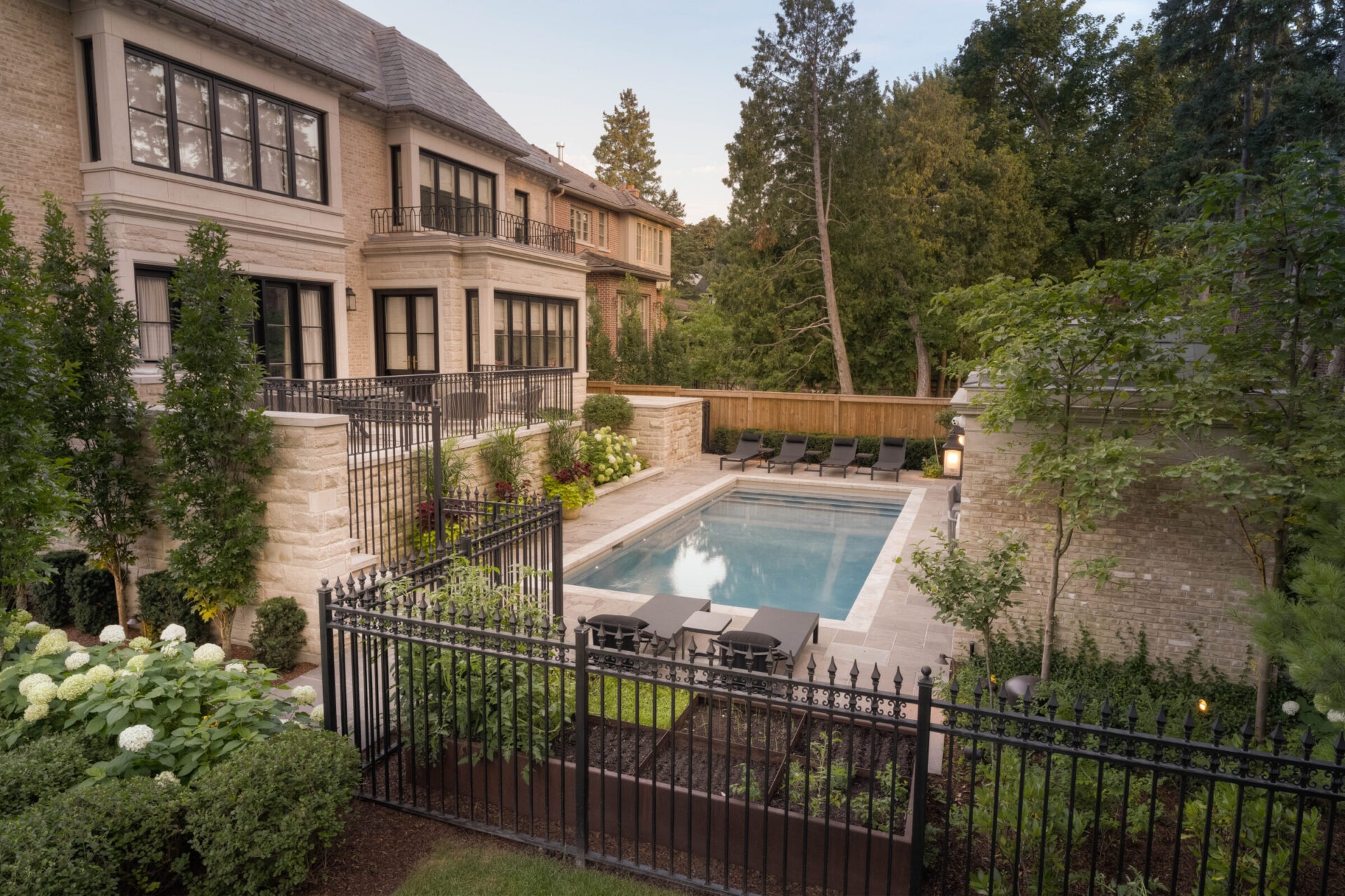 Elegant residential backyard features a rectangular swimming pool, surrounded by lush greenery and secure iron fencing, with a brick house in the background.