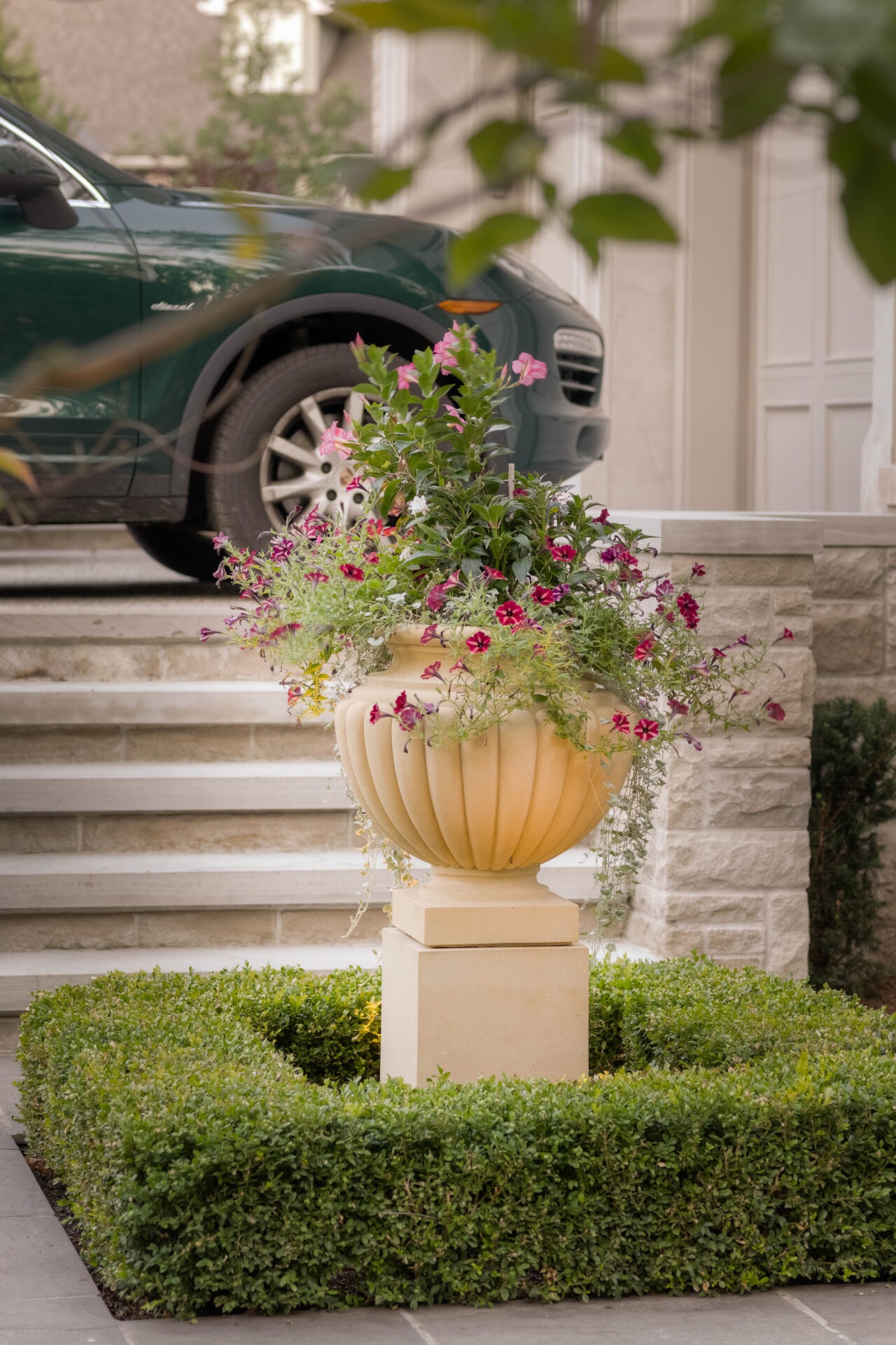 A green car parked near a staircase, with a decorative urn filled with colorful flowers, surrounded by neatly trimmed hedges.