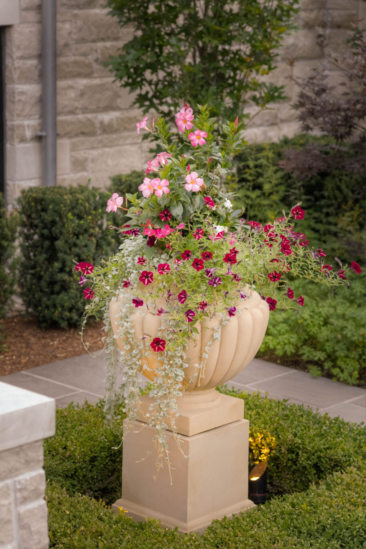 A decorative stone planter with pink and red flowers is surrounded by greenery against a stone wall backdrop in a garden setting.