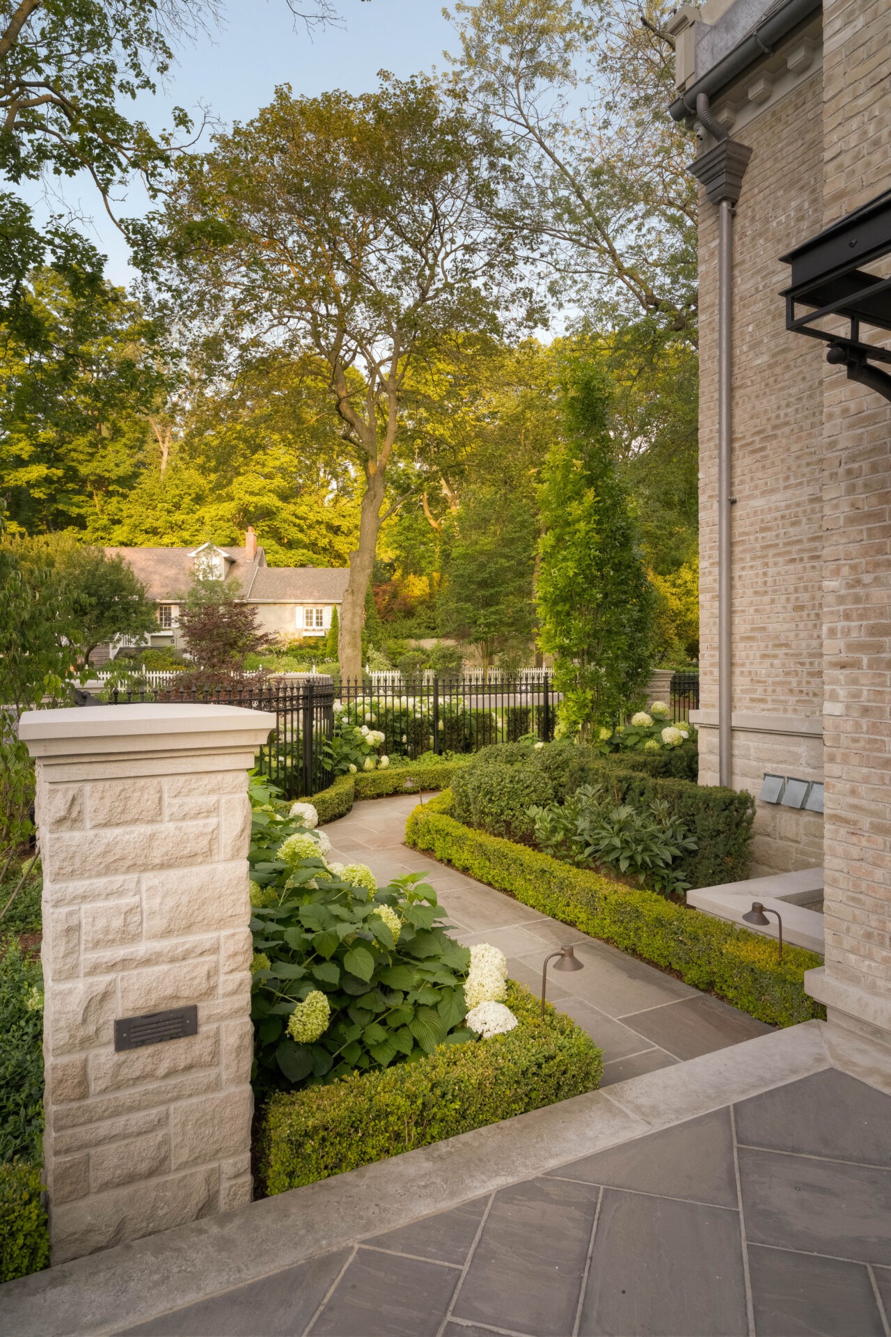 A beautifully landscaped garden with lush greenery, a stone pathway, and an elegant house exterior surrounded by trees and a picket fence.