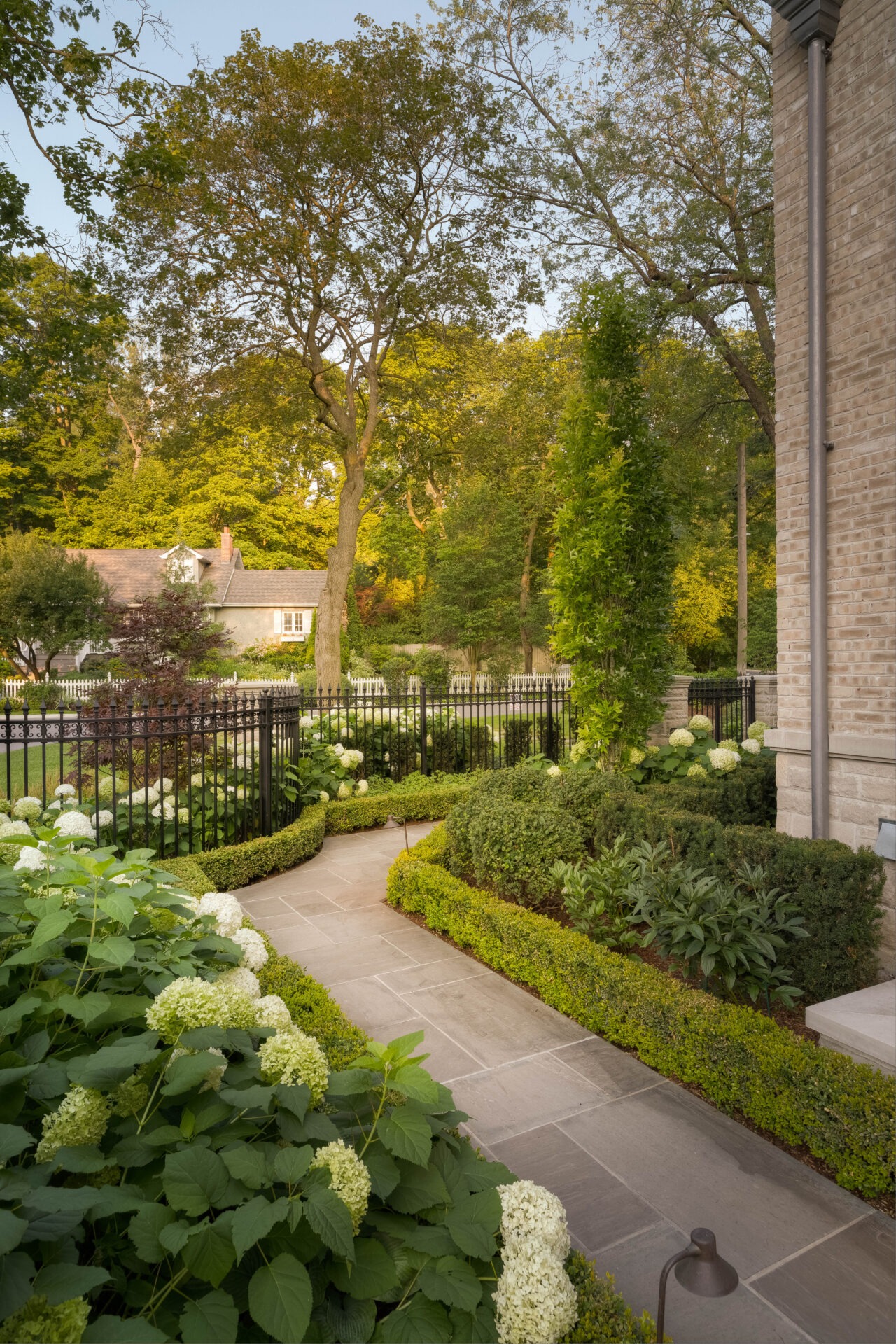 A charming garden pathway surrounded by lush greenery, hydrangeas, and trees, leading to a house with a black iron fence.