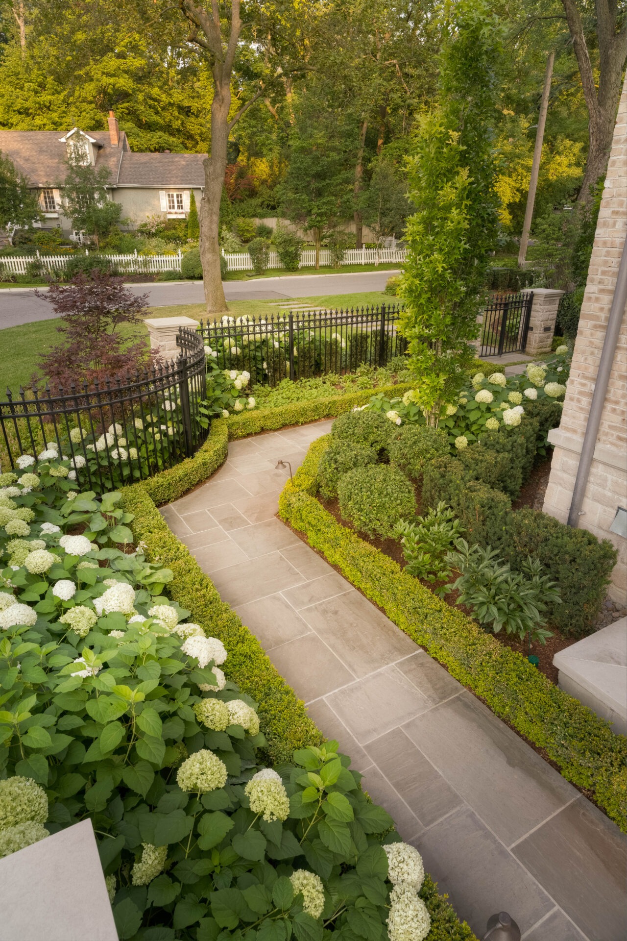 A brick path leads through a lush garden with white flowers, green hedges, and trees, towards a quaint house and picket fence.