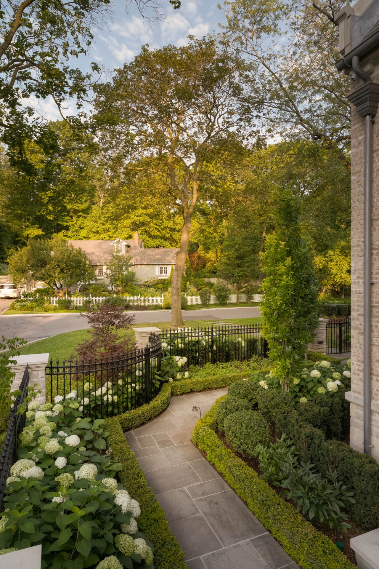 A charming house with a lush garden, stone path, and white hydrangeas. Fenced yard and trees create a serene residential scene.