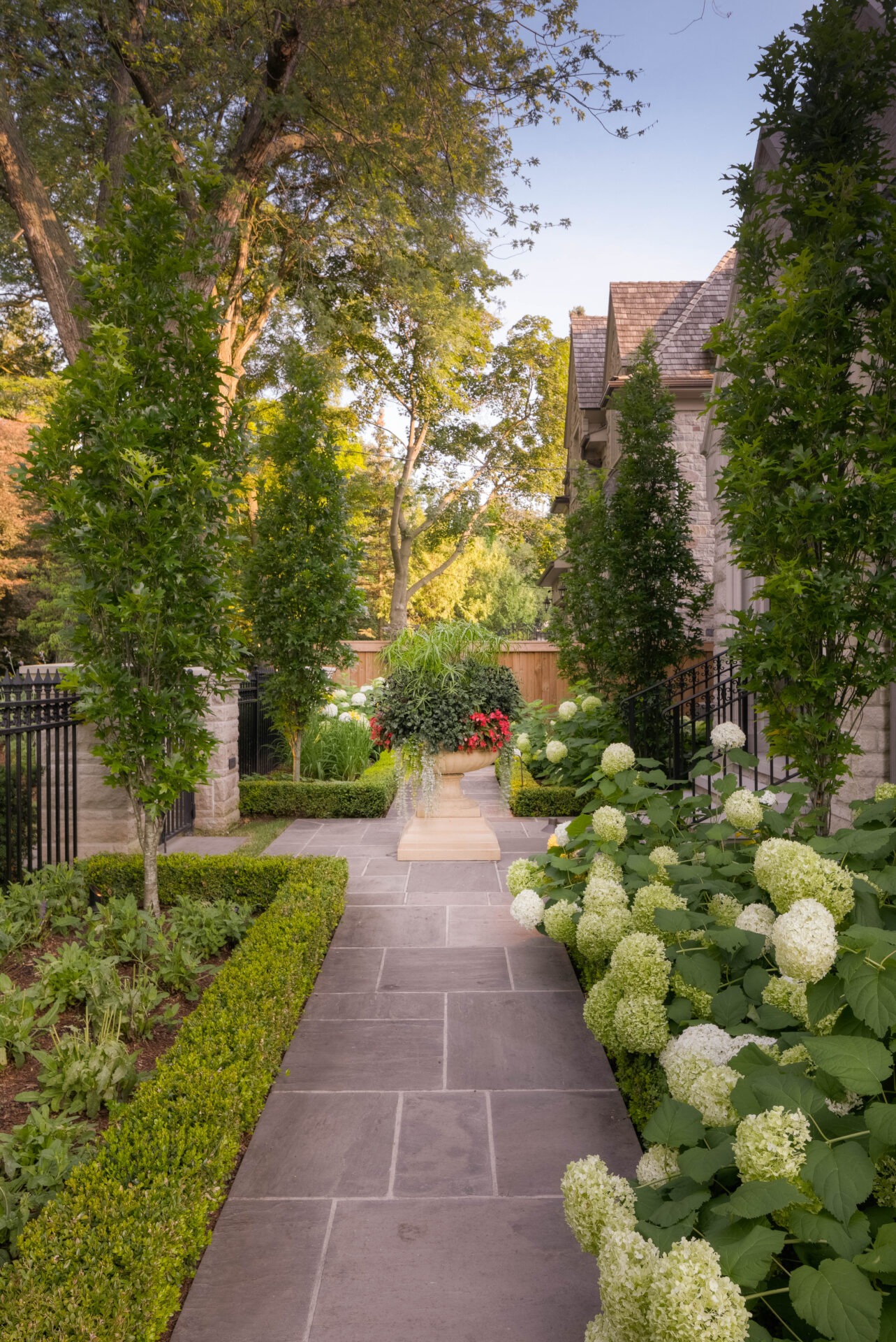 Charming garden with a stone pathway, lush hydrangeas, manicured hedges, and potted flowers, surrounded by iron fences and a brick building.