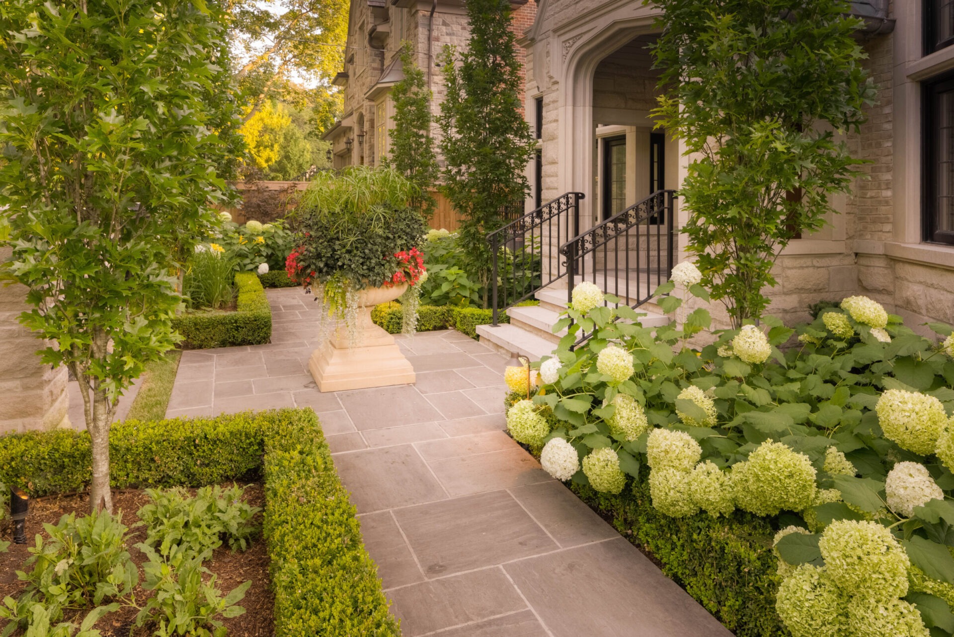 Elegant stone house front yard with manicured hedges, lush hydrangeas, and large planters. Stately entrance with steps and railings.