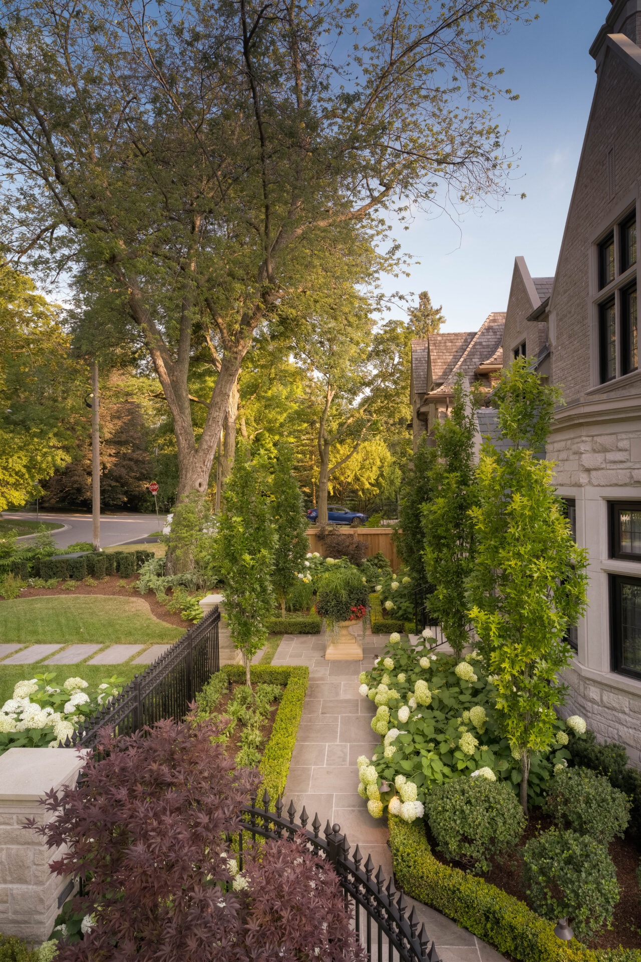 A beautifully landscaped garden with lush greenery, colorful flowers, stone pathway, and partially visible stone house on a bright, sunny day.