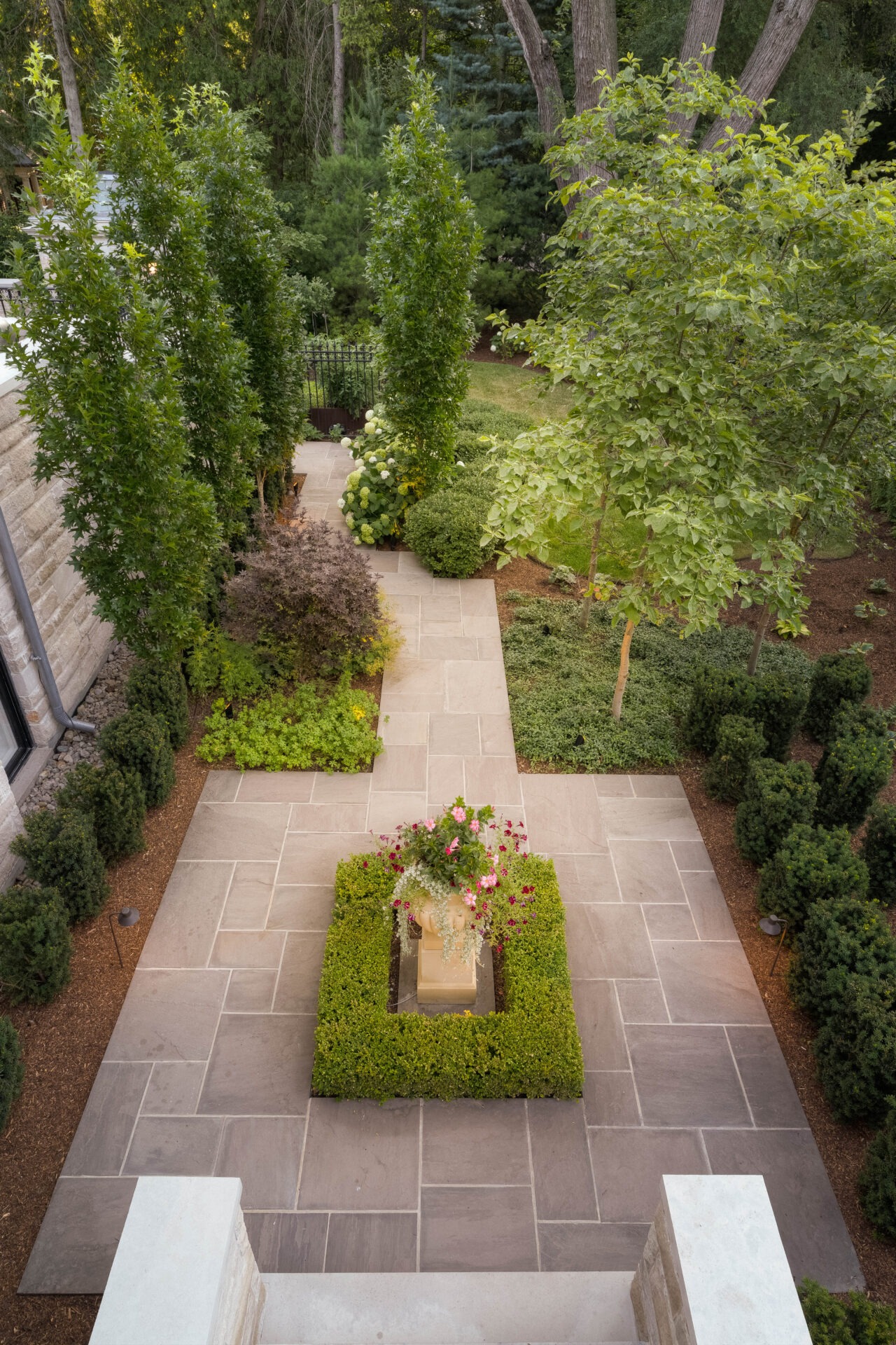 A neatly landscaped garden with paved paths, bordered by lush greenery and a central flower arrangement, surrounded by tall trees.