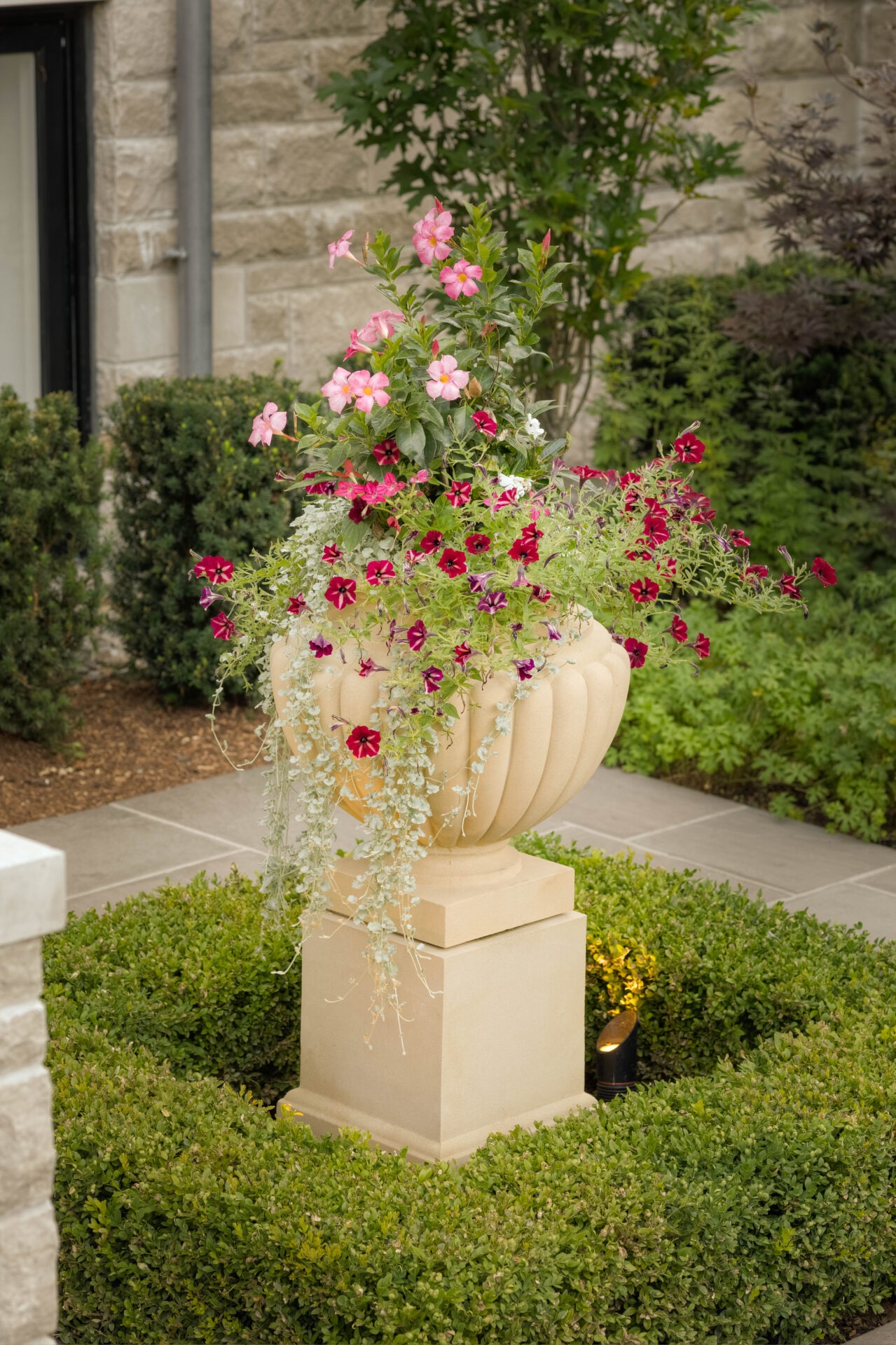 Decorative stone planter with vibrant pink and red flowers stands amid trimmed hedges and greenery, adjacent to a stone building wall.