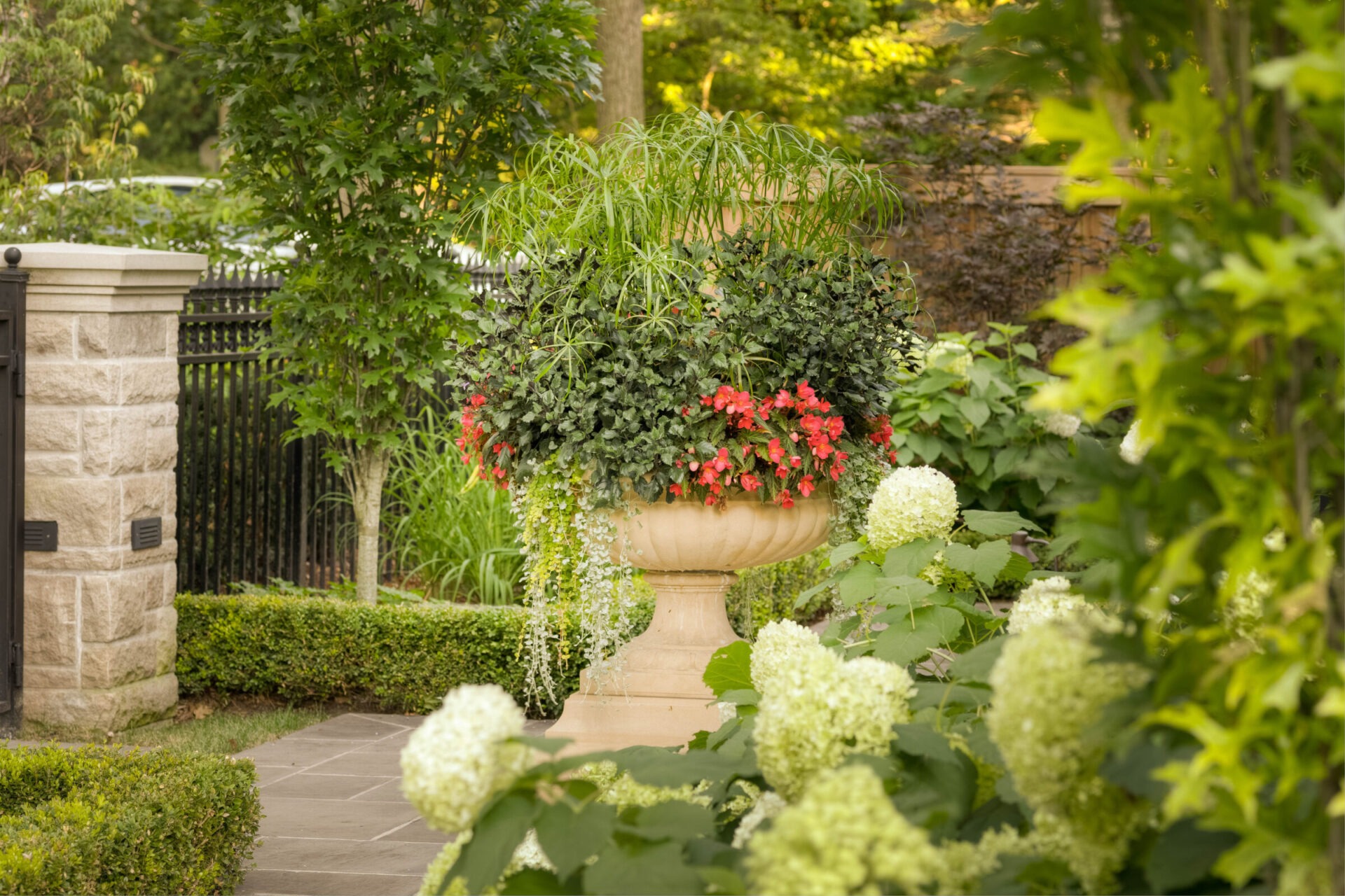 A lush garden features a large stone planter overflowing with red flowers and greenery, surrounded by hedges and hydrangeas.