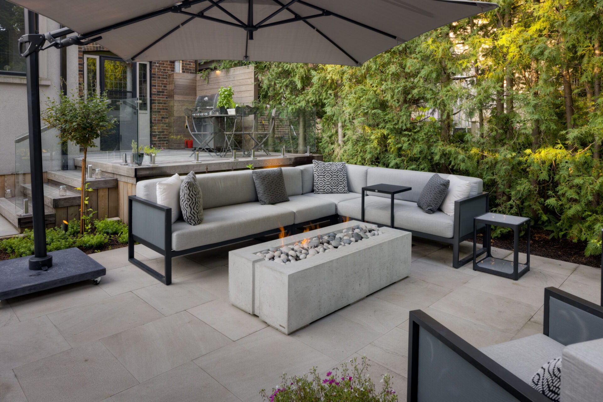 Modern outdoor patio with gray sectional sofa, fire pit table, and umbrella. Surrounded by greenery and wooden deck with grill area.