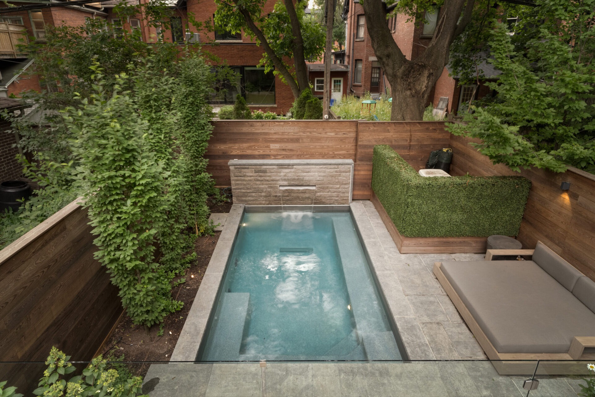 Rectangular backyard pool with waterfall feature, surrounded by wooden fencing, lush greenery, and modern seating area. Brick buildings visible in background.