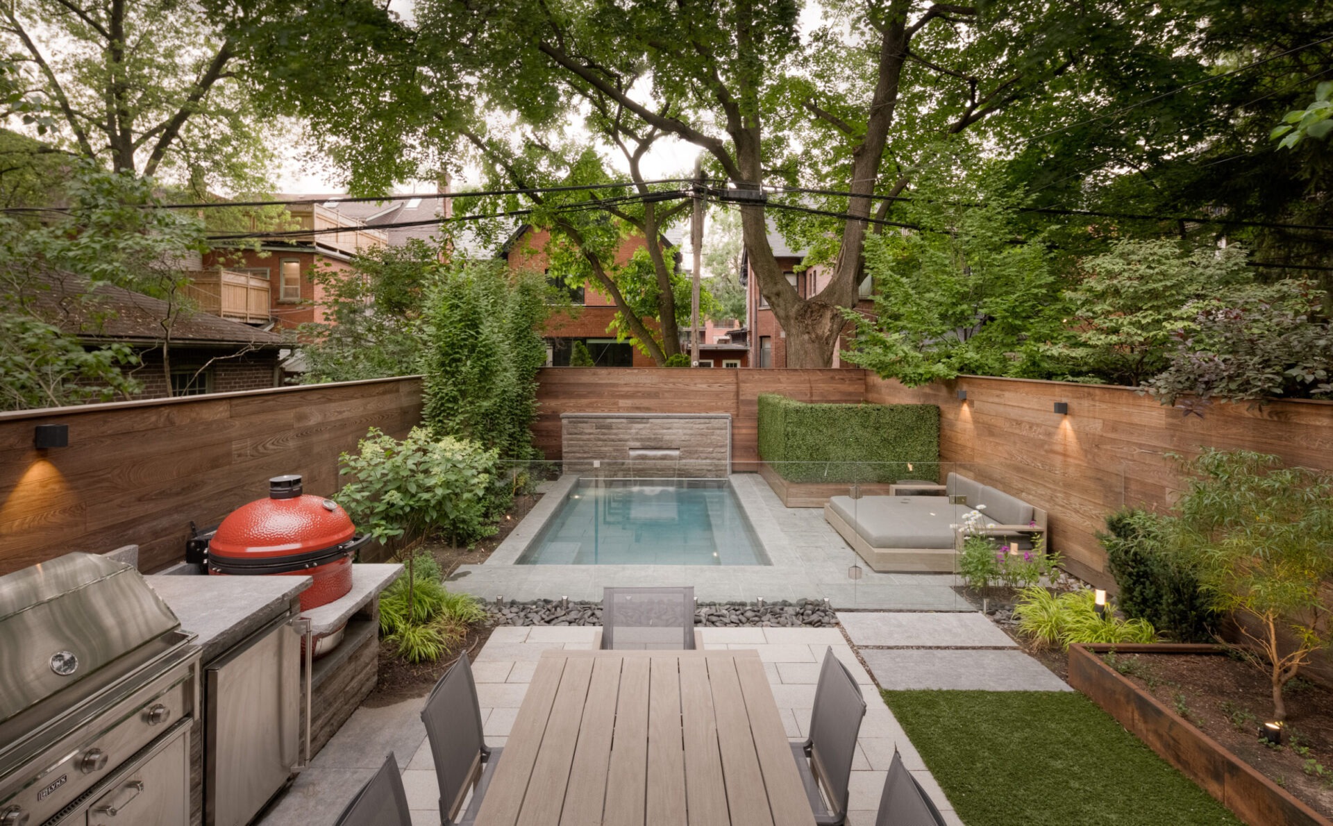 A modern backyard features a small pool, patio with seating, barbecue, and lush greenery, surrounded by wooden fencing and shaded by trees.