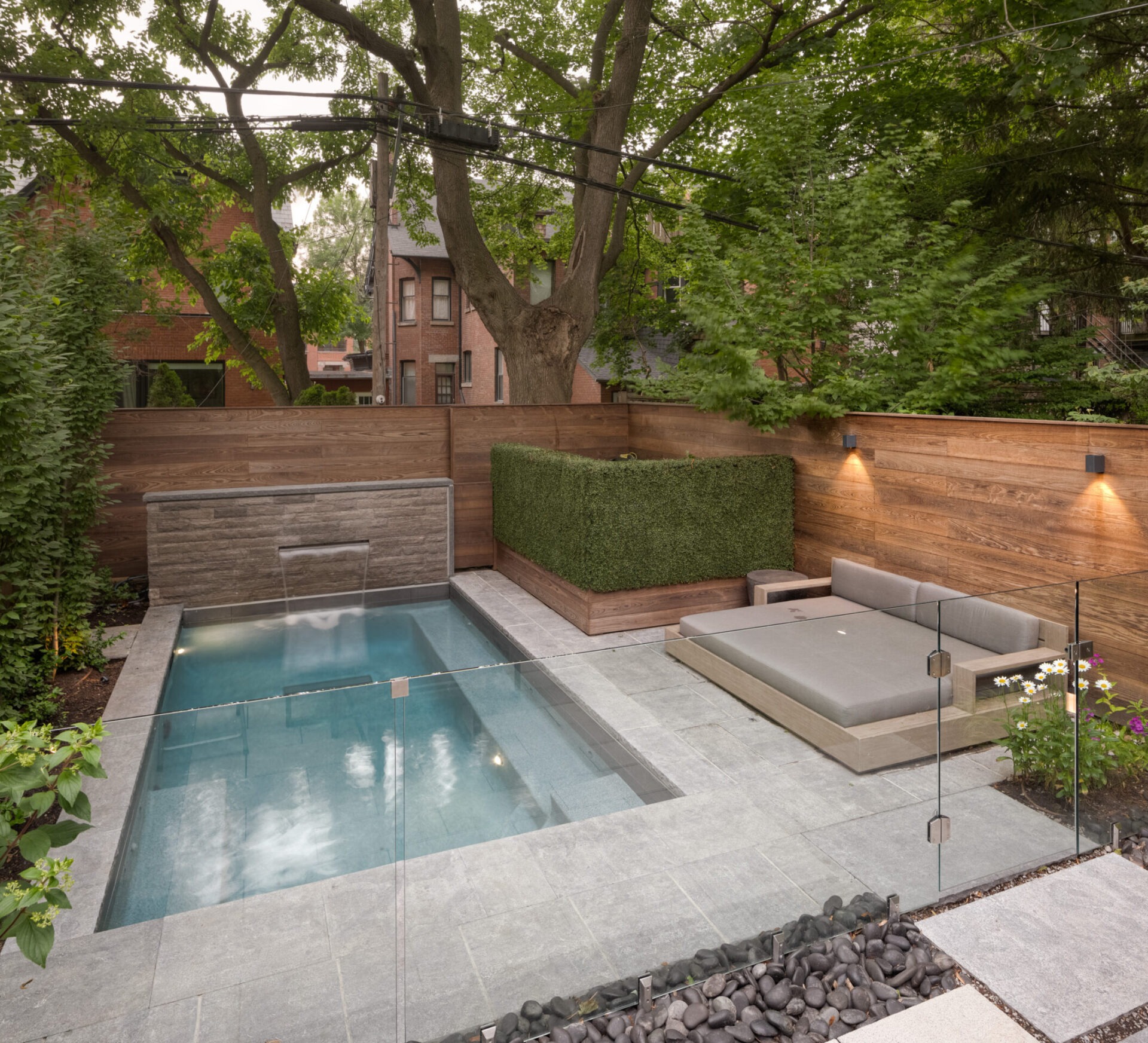 A modern backyard features a small pool with waterfall, sleek grey lounge area, wooden fencing, lush greenery, and nearby brick buildings.