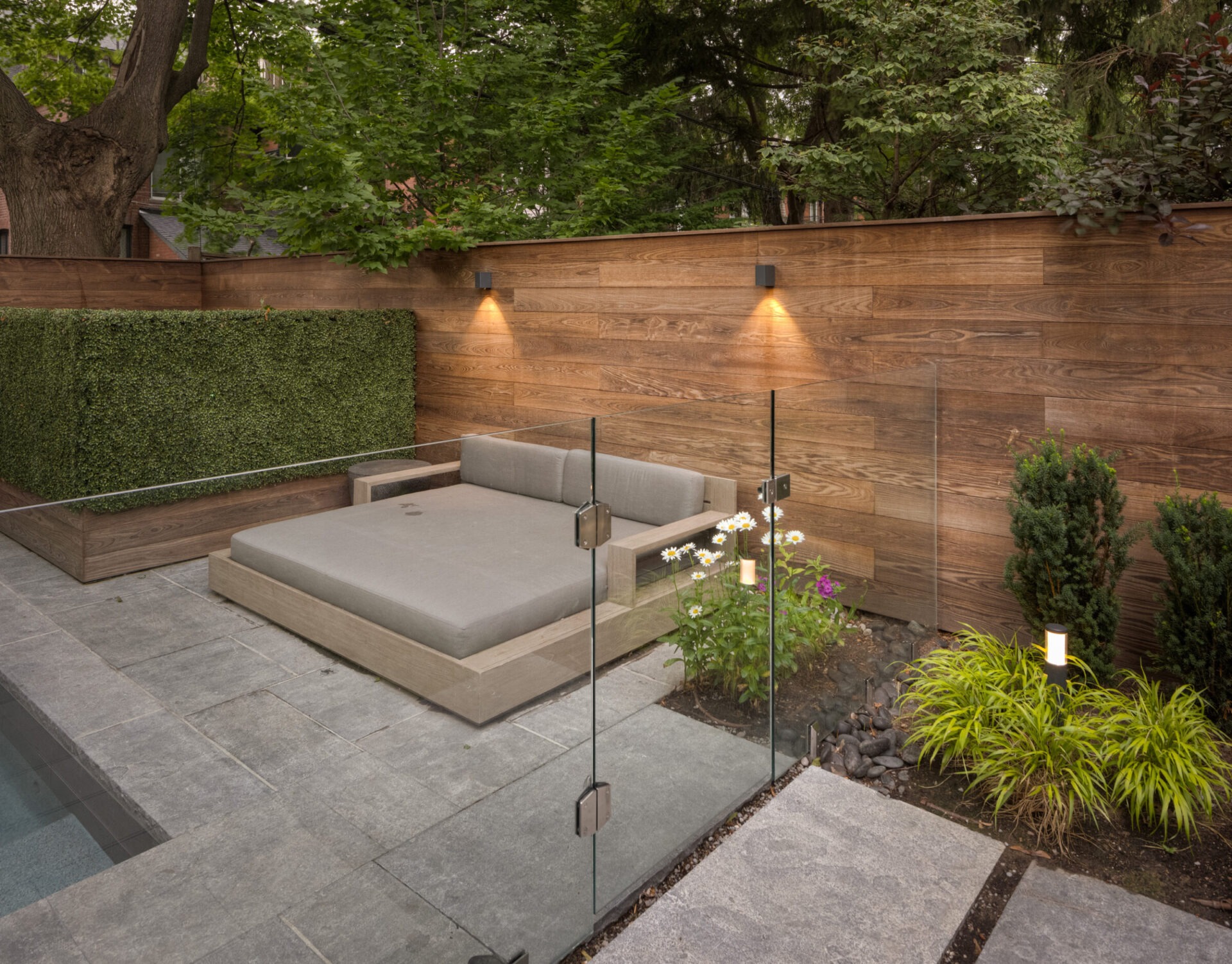 Modern backyard with wooden paneling, plants, and a gray cushioned seating area by a pool. Illuminated by warm lights, creating a serene atmosphere.