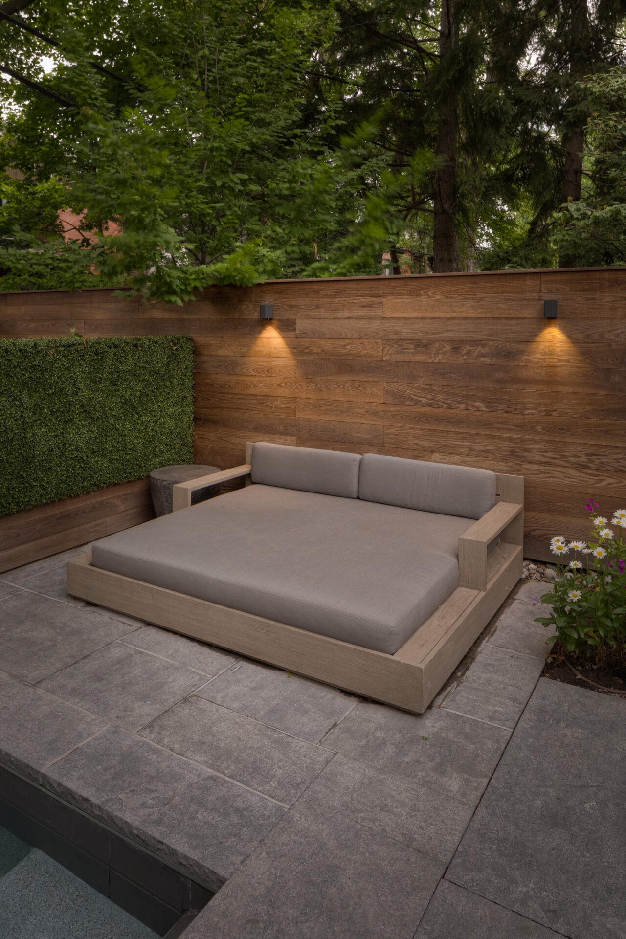 Outdoor patio with a large wooden daybed, surrounded by greenery and illuminated by wall lights, creates a serene, cozy atmosphere.