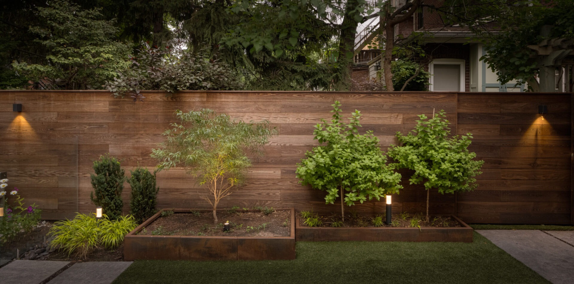 A serene garden with wooden fence, lush greenery, and soft lighting, creating a tranquil outdoor space beside a residential building.