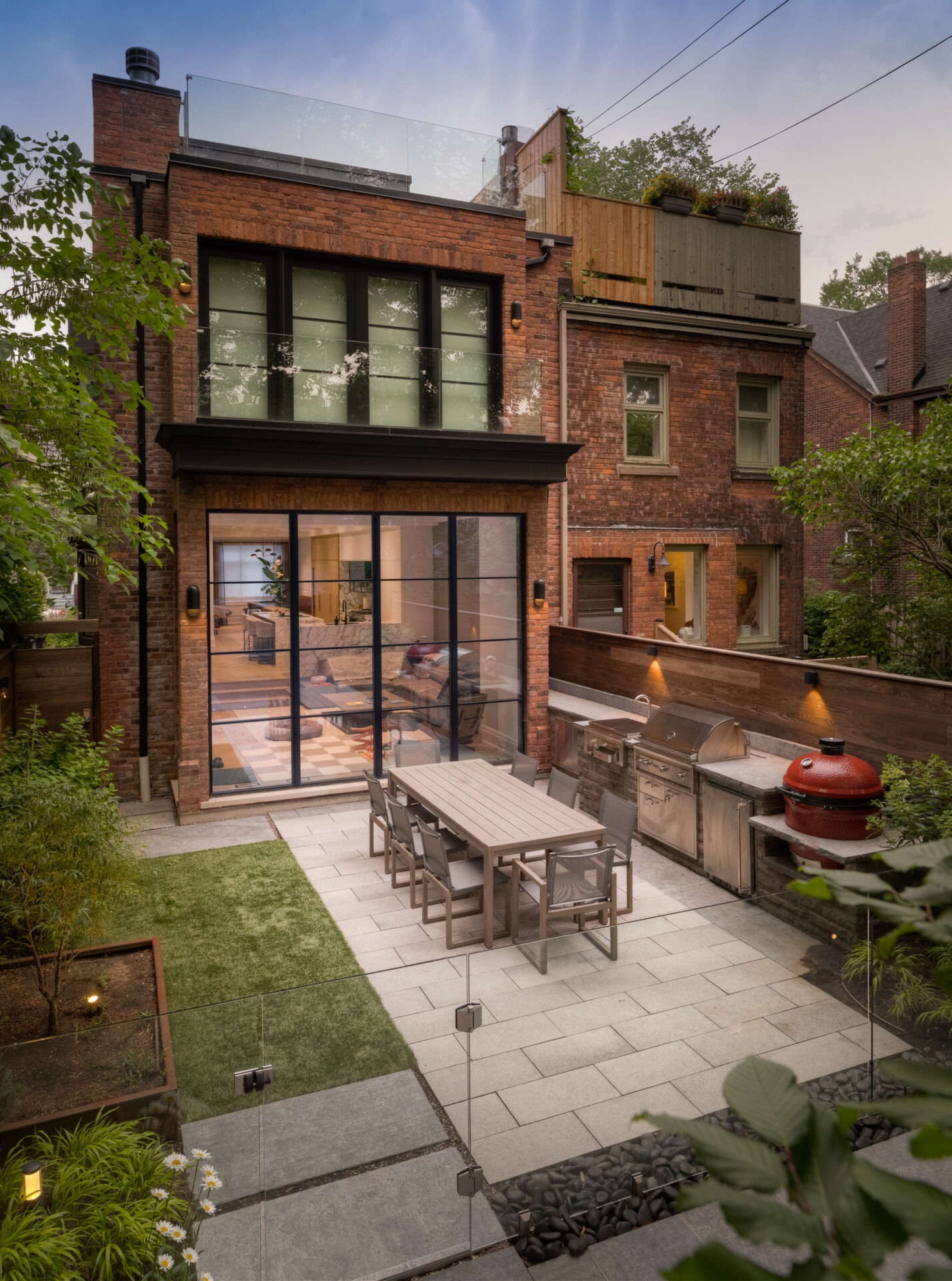 Modern brick house with glass doors opens to a patio featuring a dining table, grill, and lush greenery. Cozy and inviting outdoor space.