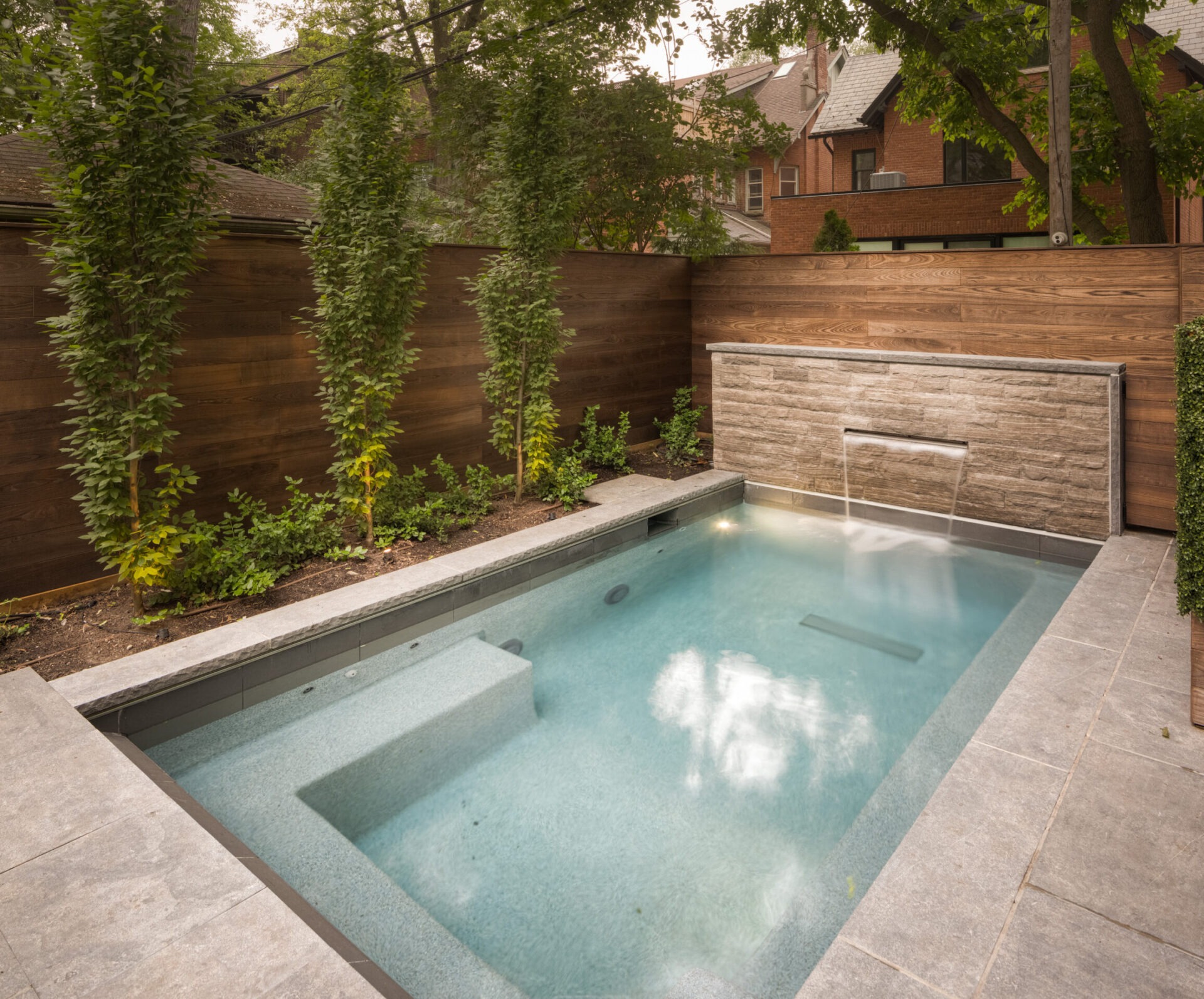 A backyard with a small pool features a stone waterfall. Tall trees and wooden fencing enhance the serene, private setting.