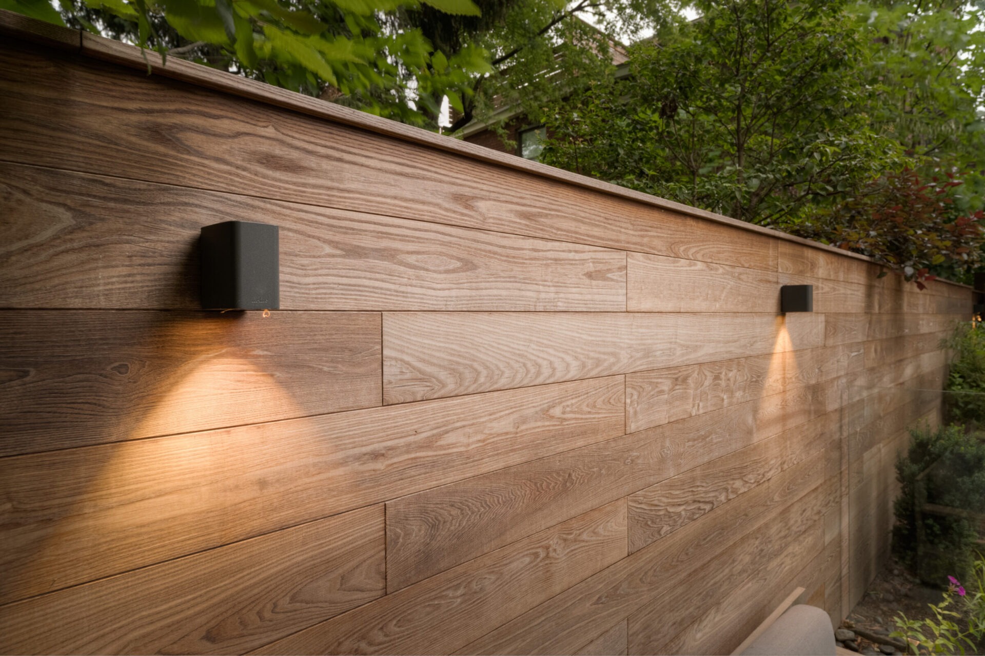 Wooden fence with integrated lights illuminating downwards, surrounded by lush greenery. The modern, sleek design complements the natural setting.