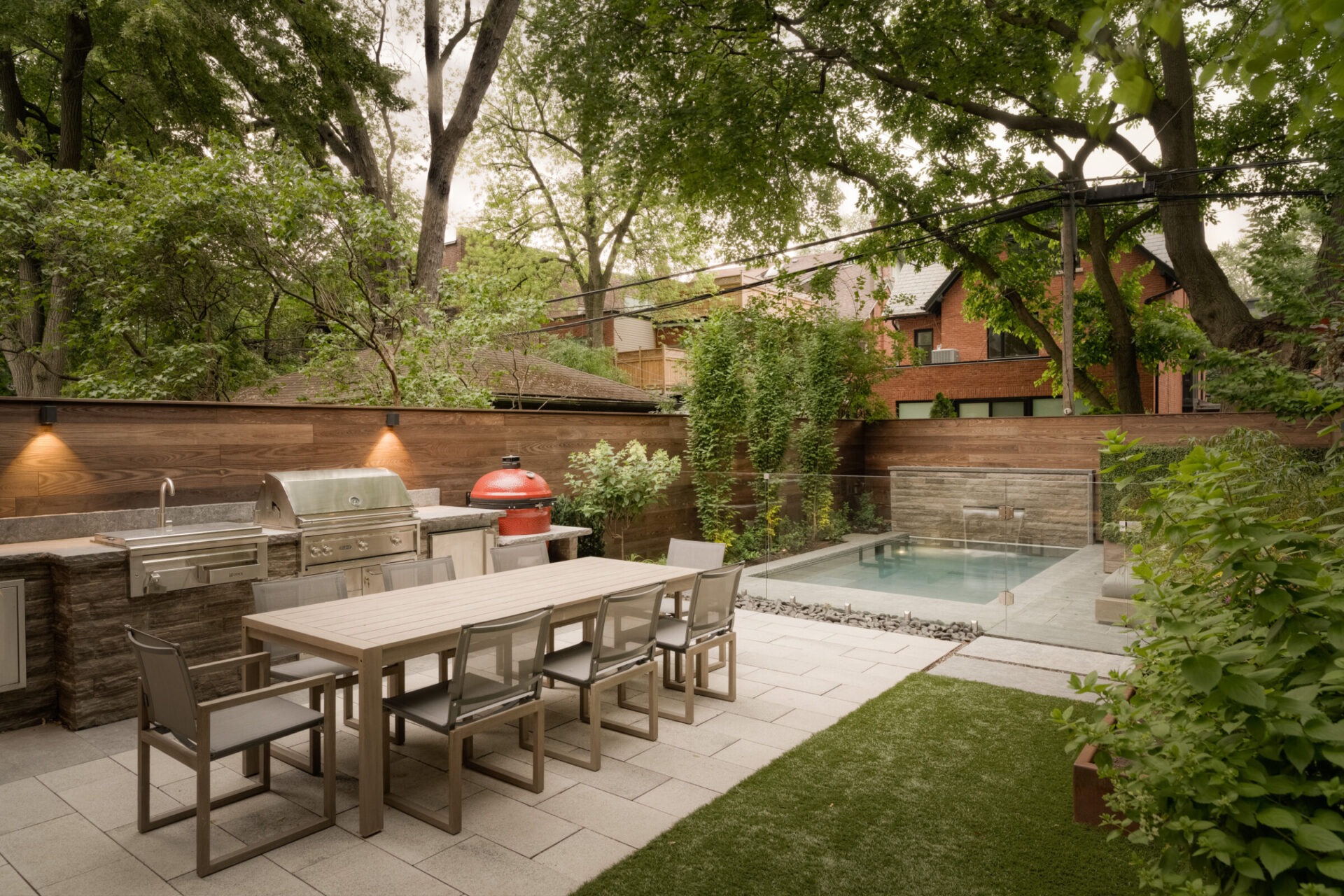 A modern backyard features a dining table, barbecue area, pool, and lush greenery, enclosed by wooden fencing and surrounded by tall trees.