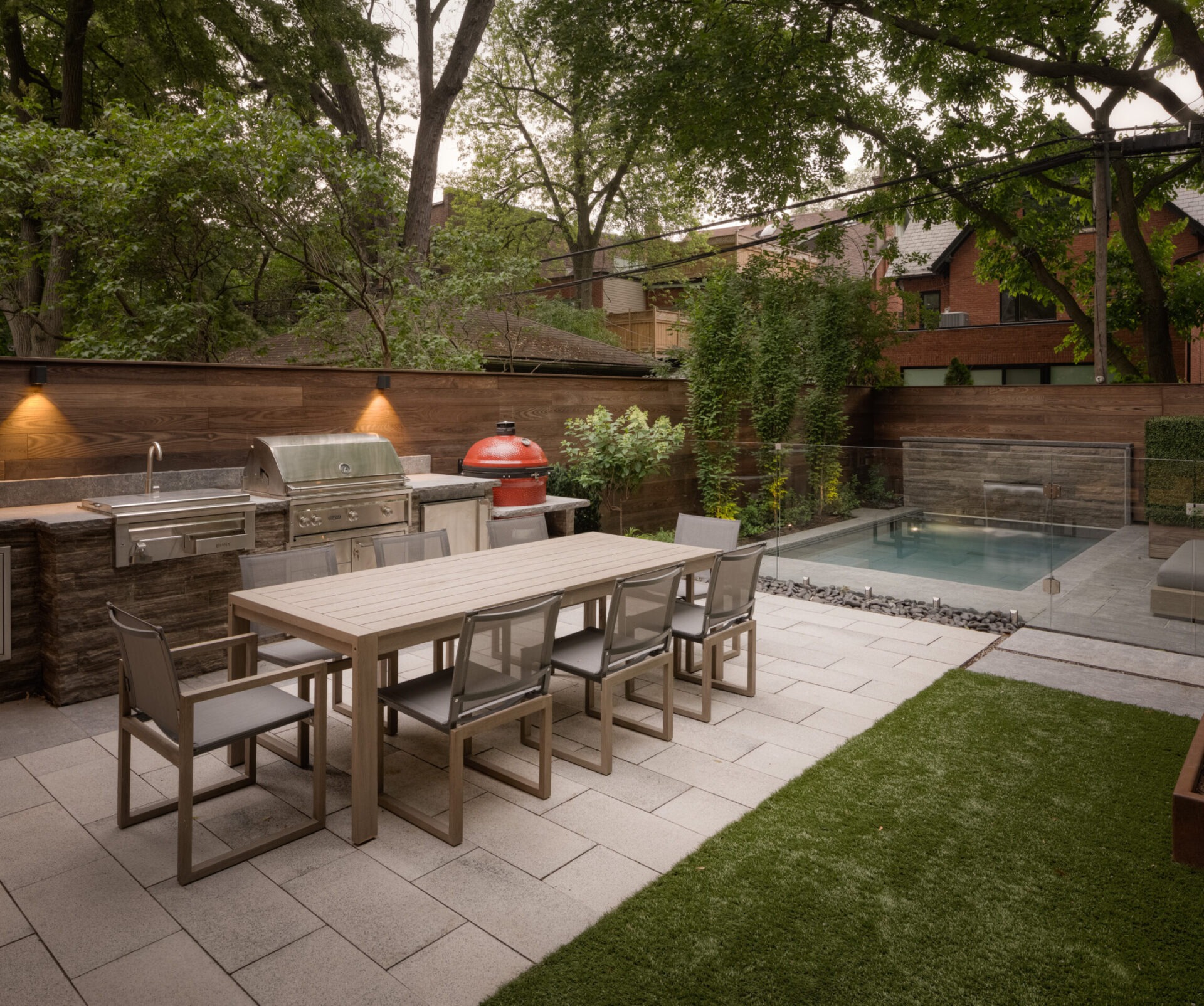 A modern backyard featuring an outdoor kitchen, dining table, and small pool surrounded by greenery and a wooden privacy fence.