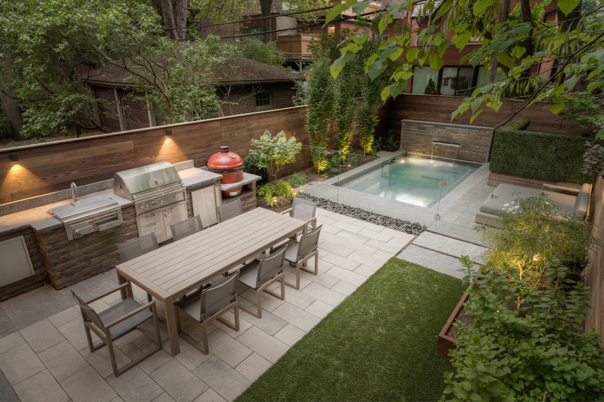 Modern backyard with a dining table, built-in grill, small pool, lush greenery, and ambient lighting, creating a cozy outdoor living space.