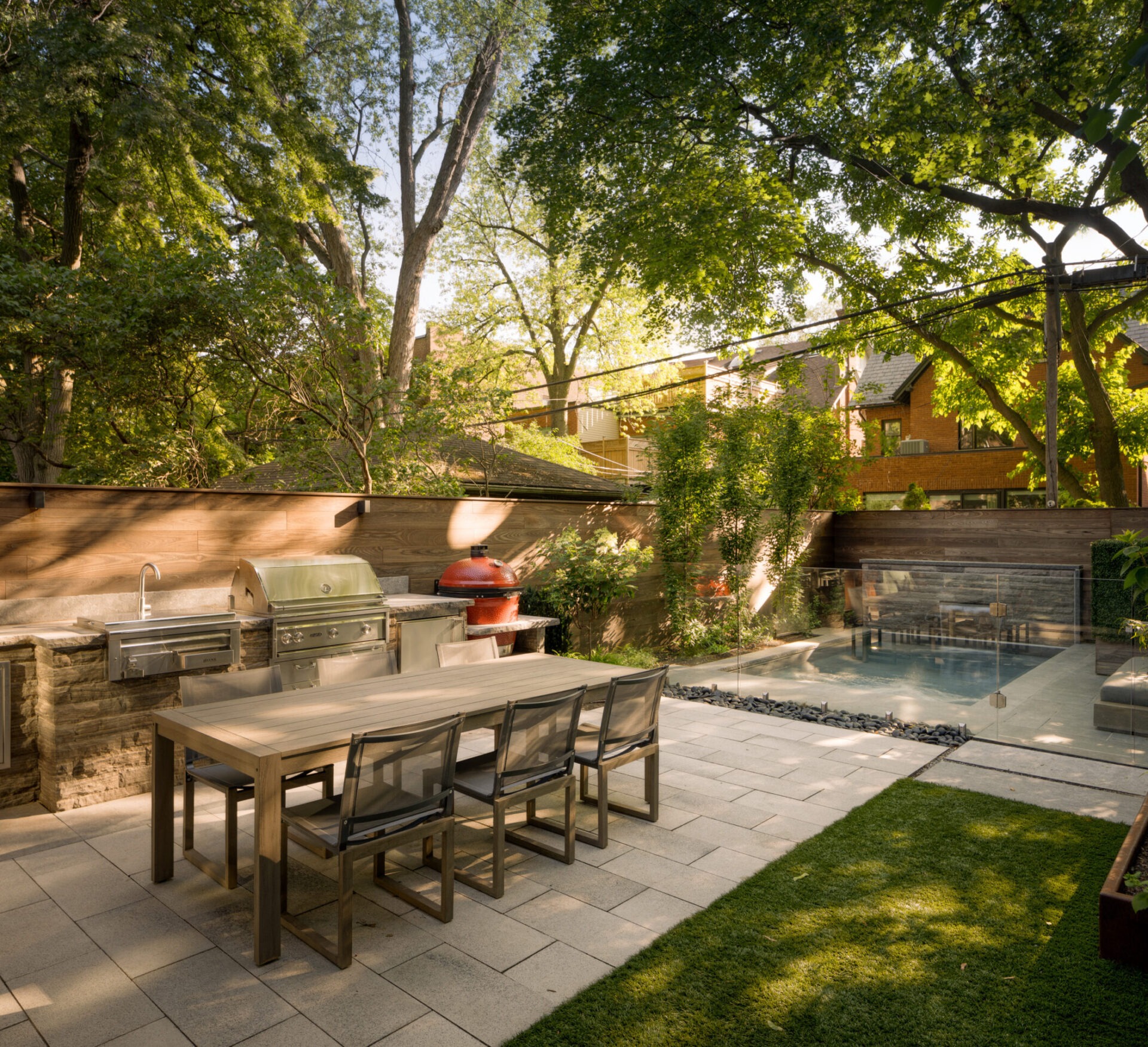 A modern backyard features a dining area with grill, surrounded by lush trees, patio, and a small pool with glass barrier.