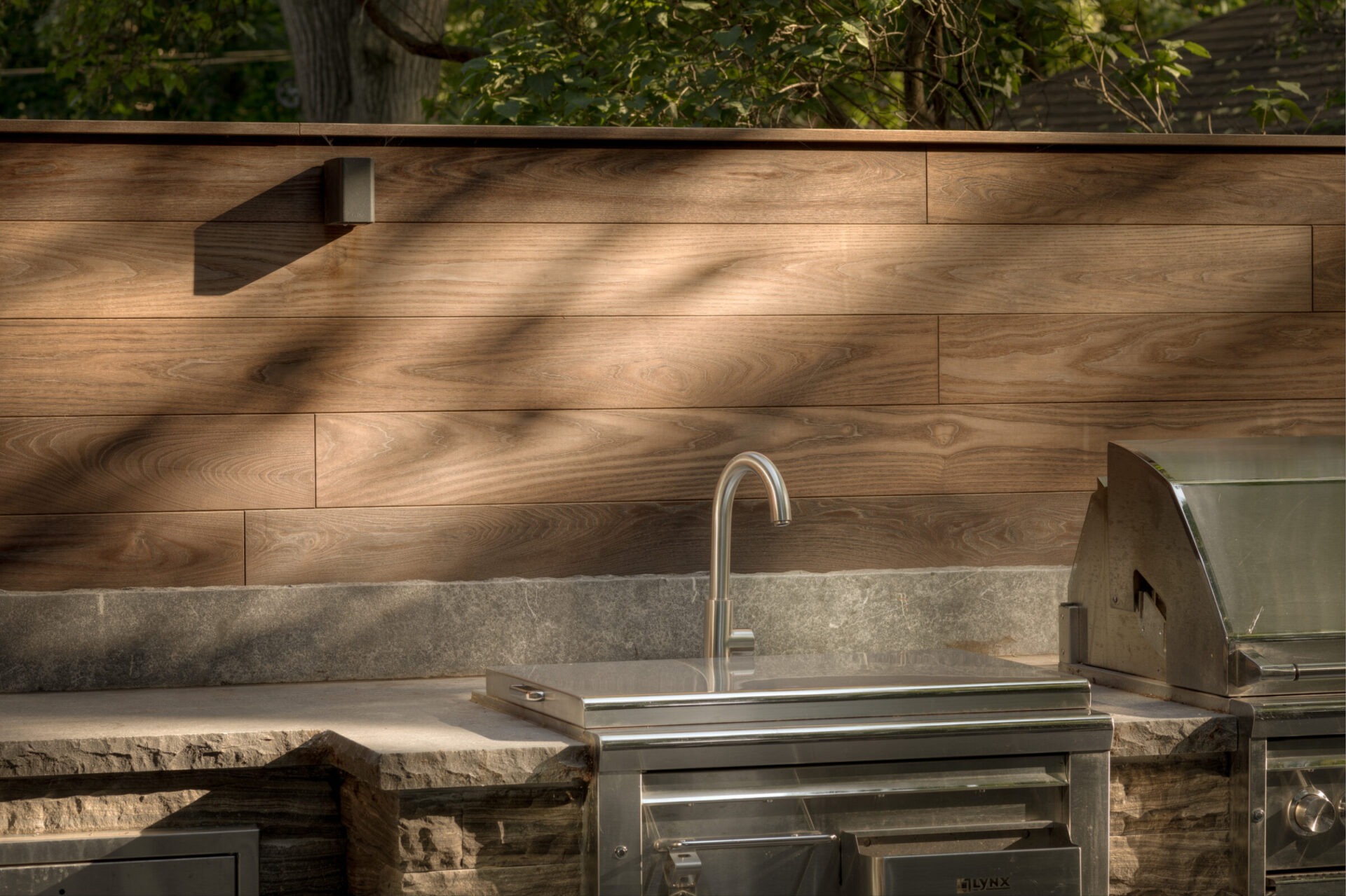 Outdoor kitchen with wooden backsplash, stainless steel sink, and grill under dappled sunlight and surrounded by greenery. Tranquil and inviting setting.