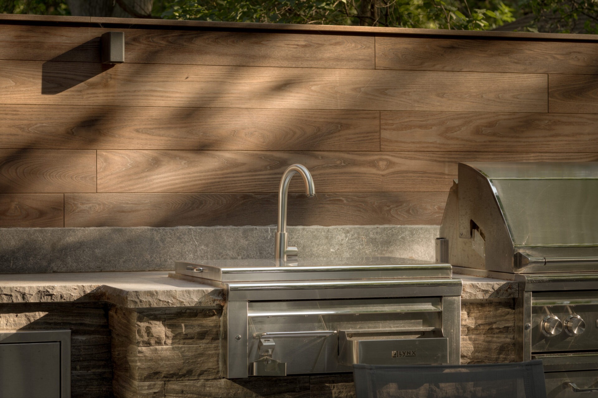 An outdoor kitchen features stainless steel appliances, a sleek metal faucet, and a wooden wall backdrop under dappled sunlight, framed by greenery.