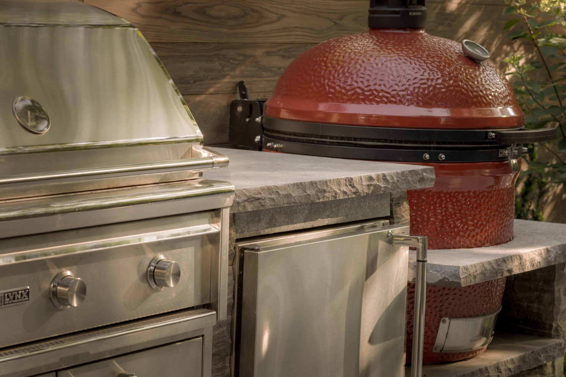 Outdoor kitchen with a stainless steel grill and a red ceramic kamado smoker on a stone counter, surrounded by greenery.