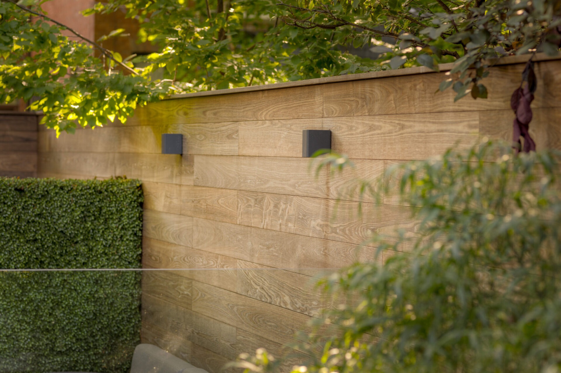 Wooden fence with two lights, surrounded by lush green foliage and bushes, creating a serene garden atmosphere with dappled sunlight.