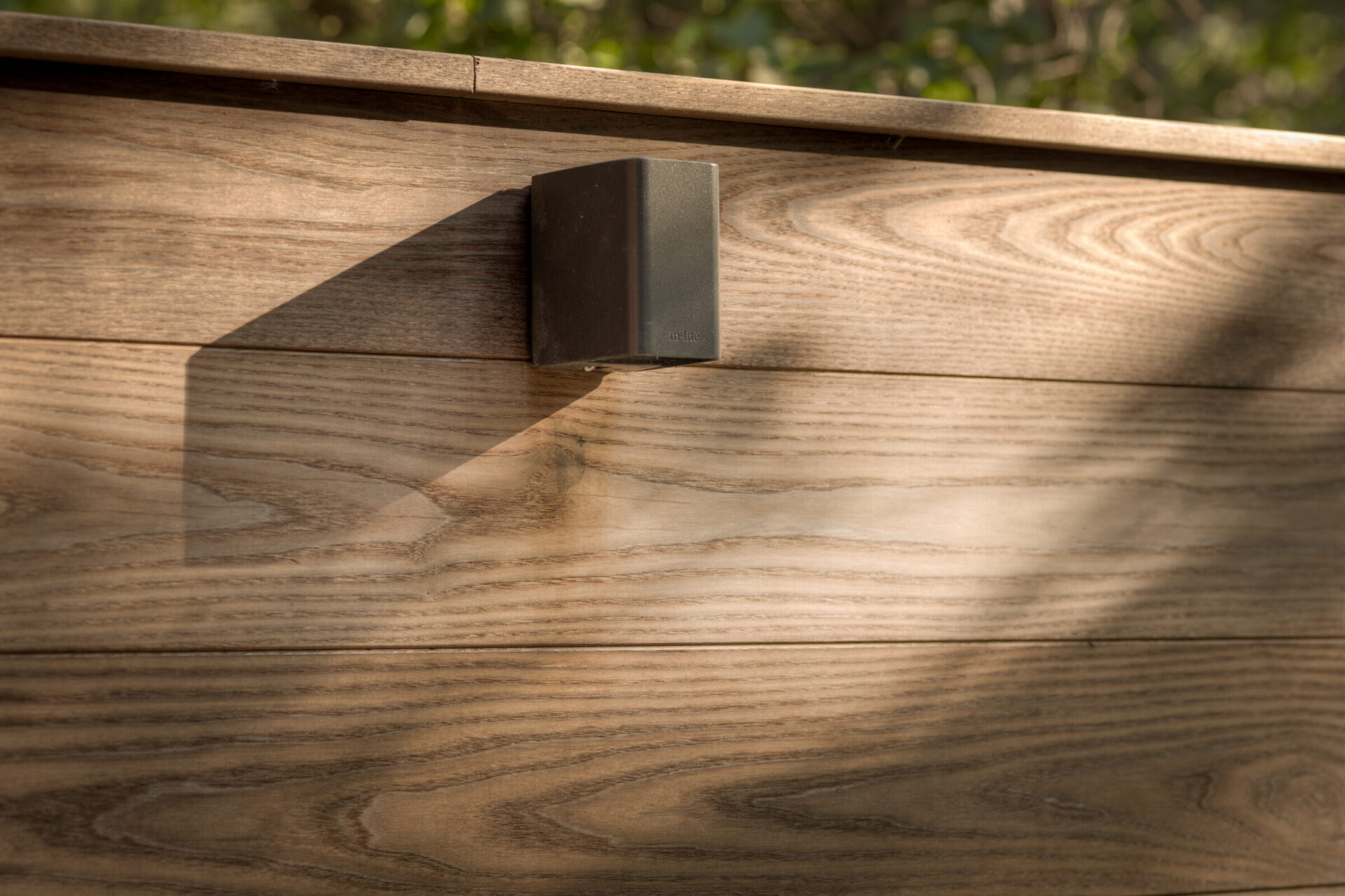 Wooden wall with a small, dark rectangular fixture casting a long shadow in sunlight. Simple, natural outdoor setting.