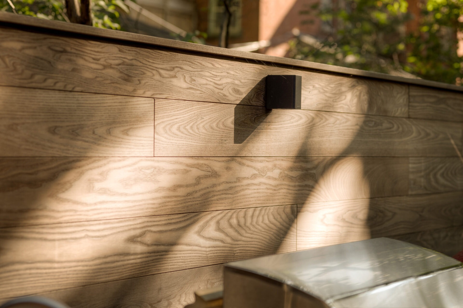 Wooden deck with a shadow from a tree, featuring a black wall lamp under sunlight. Appears to be a residential area.