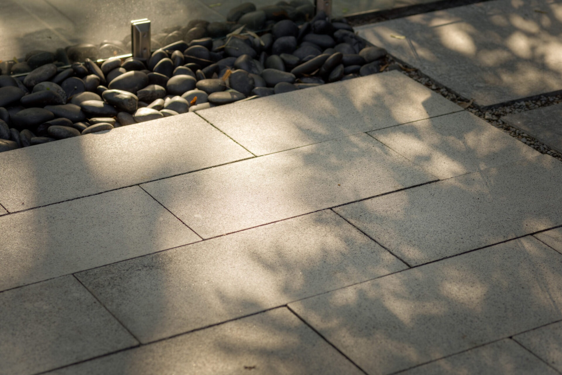 Sunlit concrete pavement with leaf shadows, bordered by smooth black stones and a glass barrier. A reflective, serene outdoor scene.