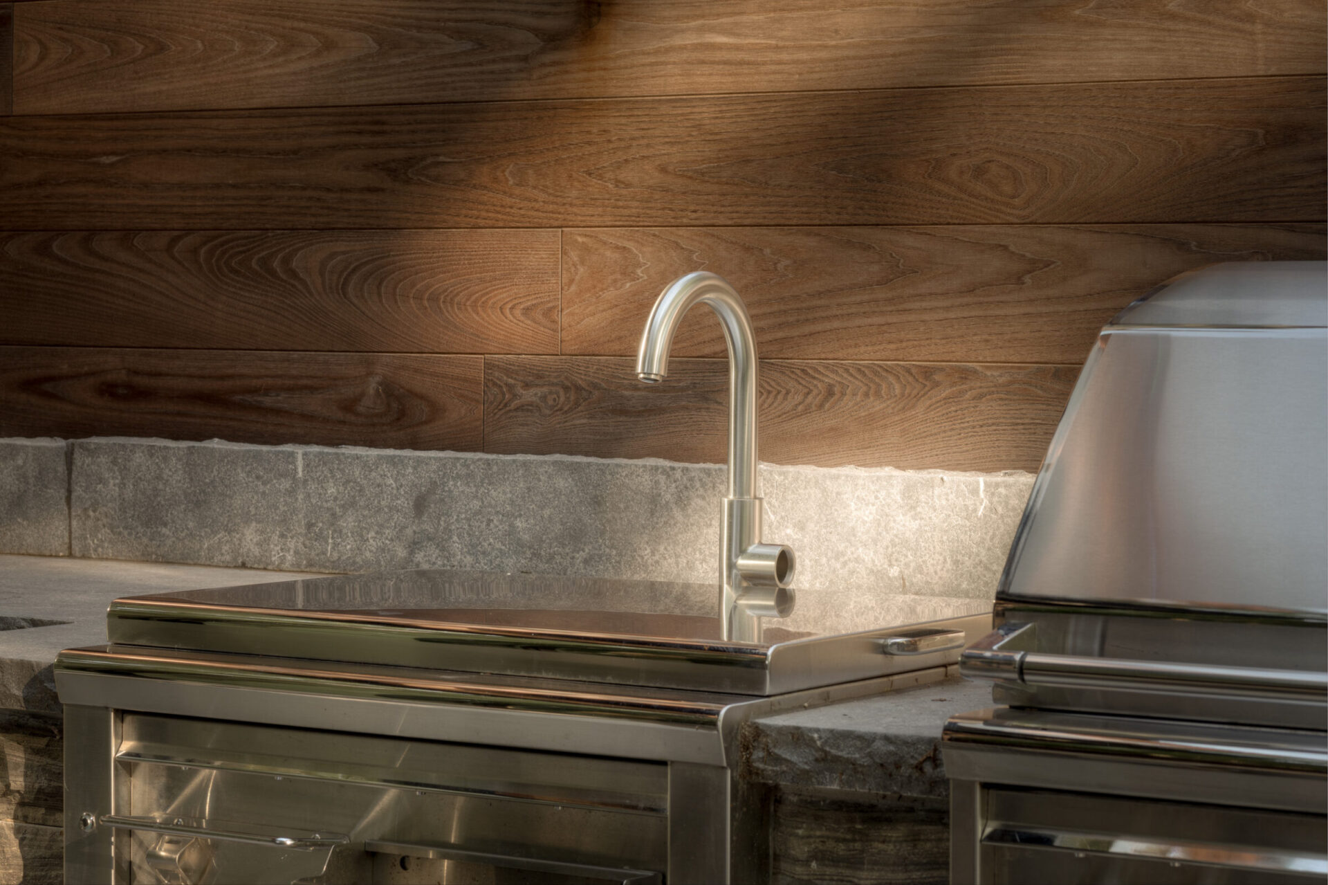 Outdoor kitchen with stainless steel fixtures, grill, and stone countertop against a wooden wall. Modern and sleek design for culinary activities.