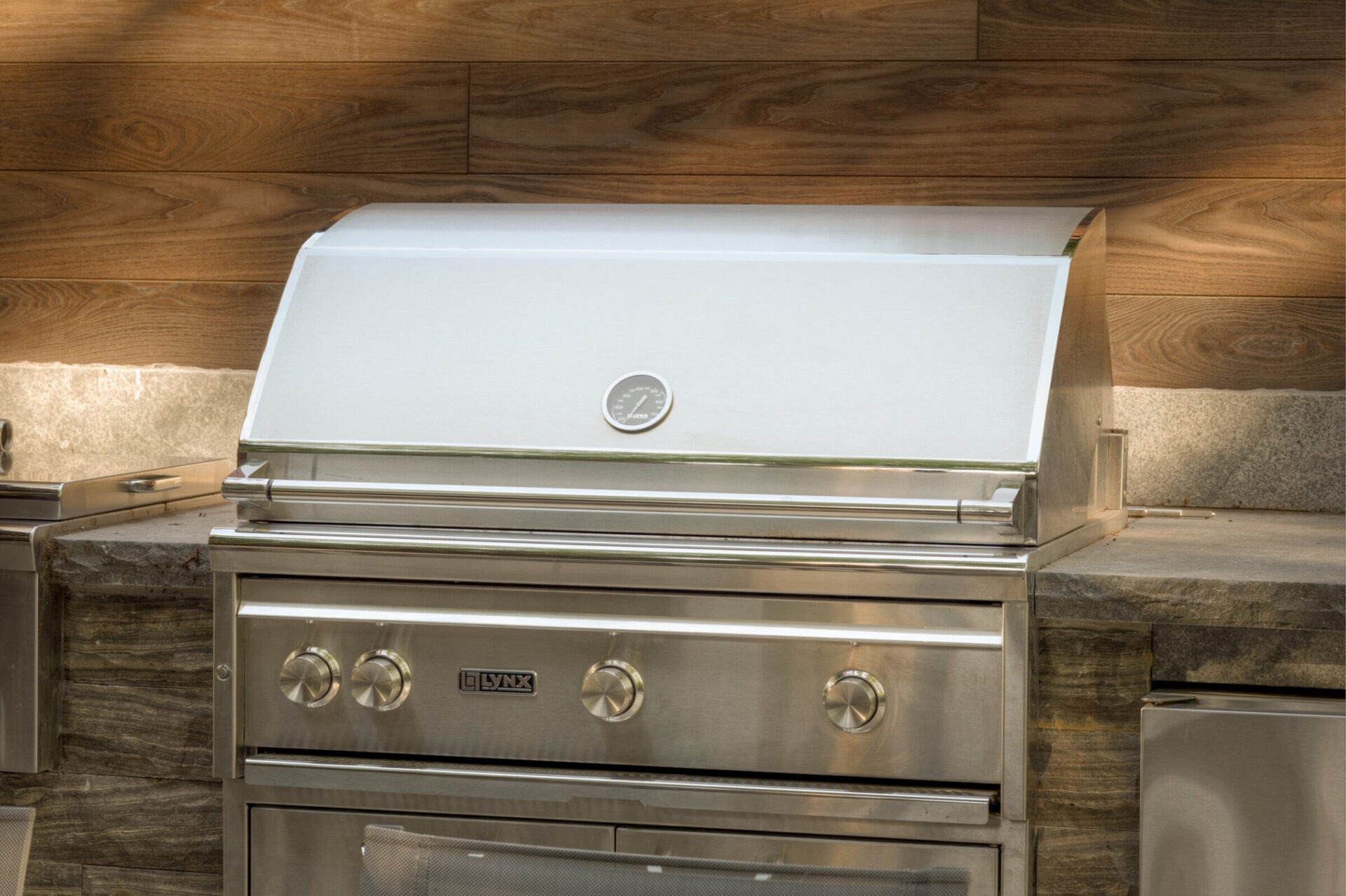 A modern stainless steel grill with closed lid sits on a stone countertop, set against a wooden paneled wall backdrop.