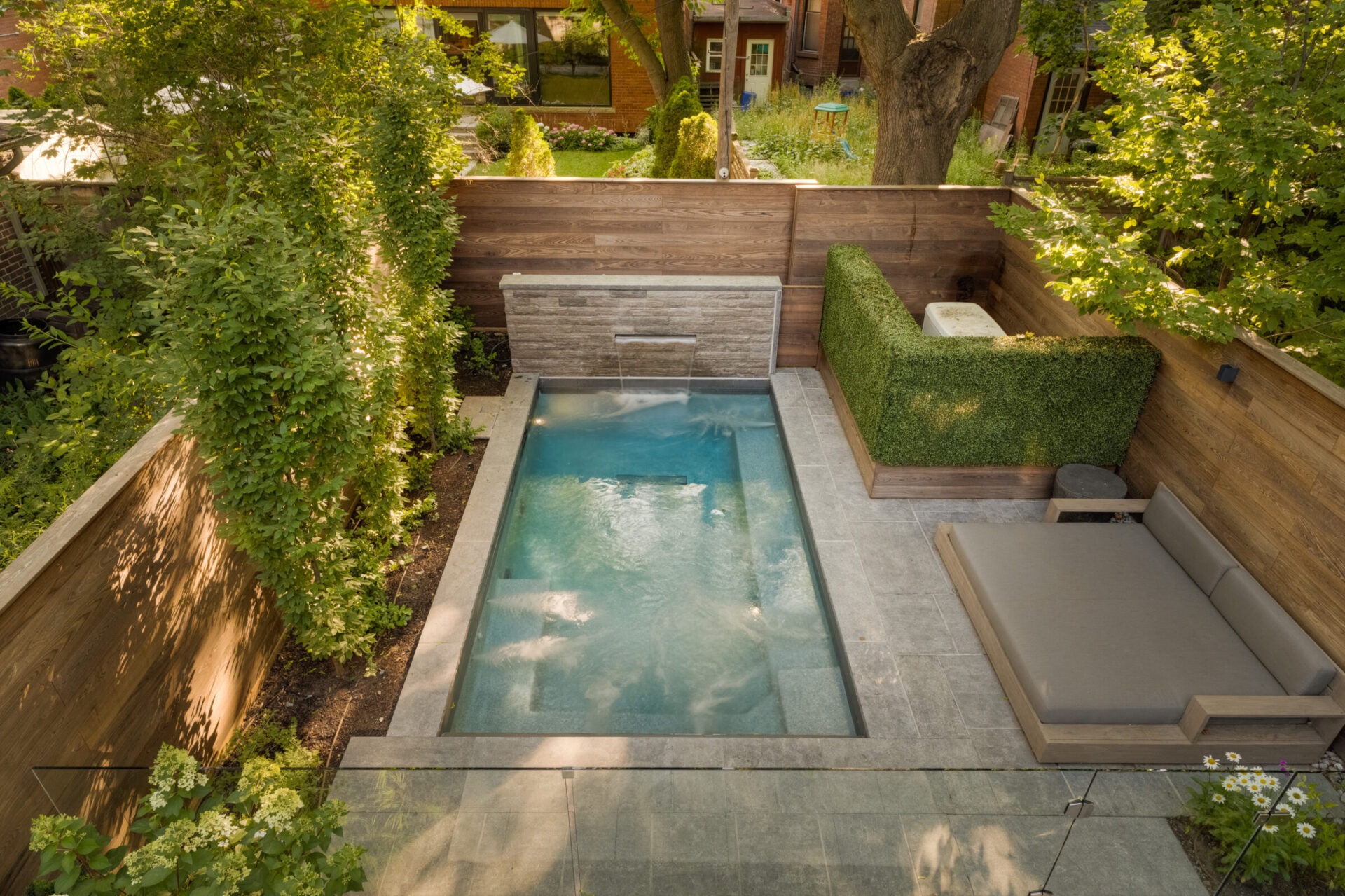 A small, rectangular backyard pool surrounded by lush greenery, wooden fence, and gray tile floor, features a nearby gray outdoor sofa.