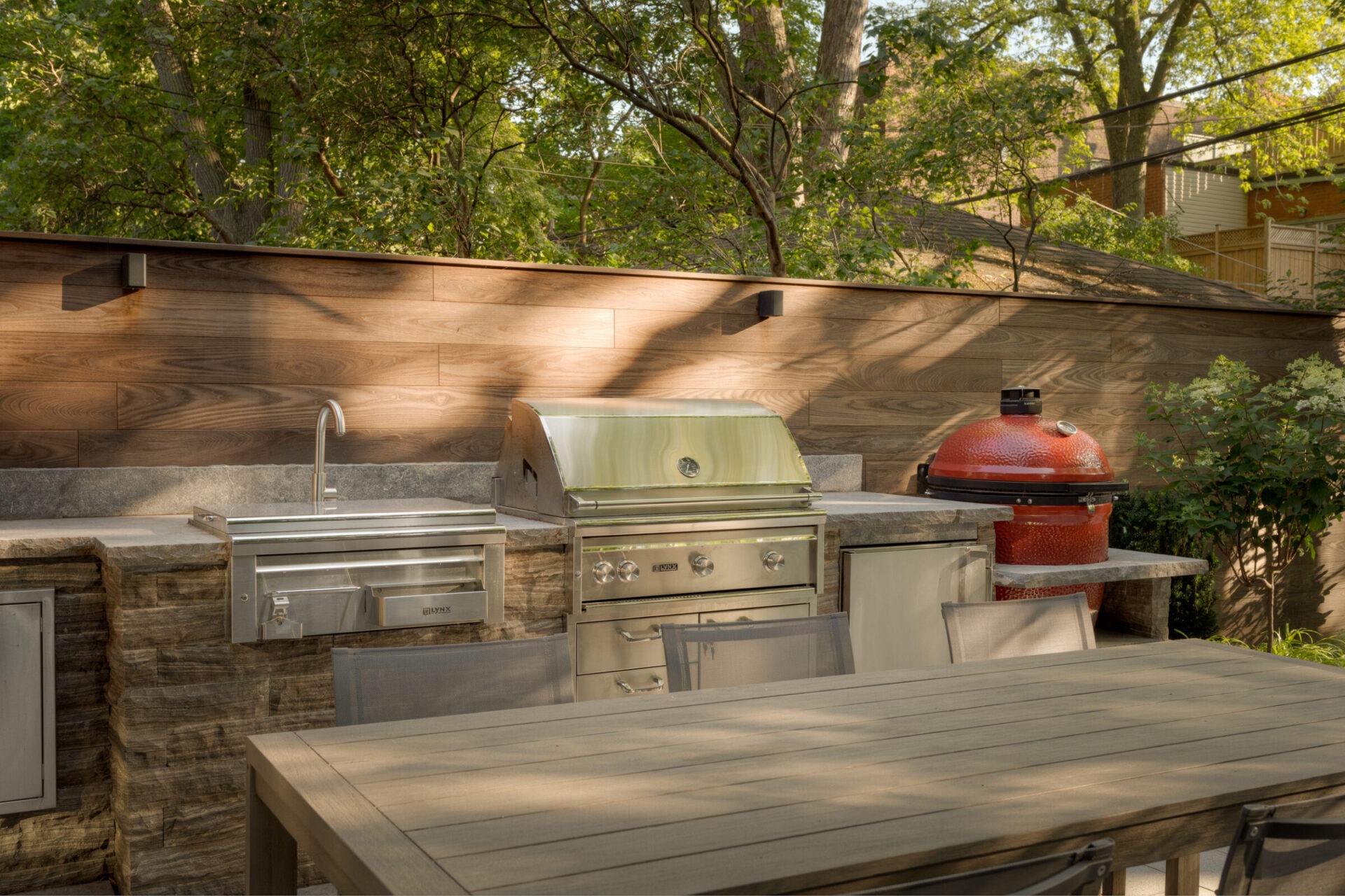 Outdoor kitchen with metal grill, red ceramic smoker, and wooden table surrounded by trees, creating a serene, natural atmosphere in the backyard.