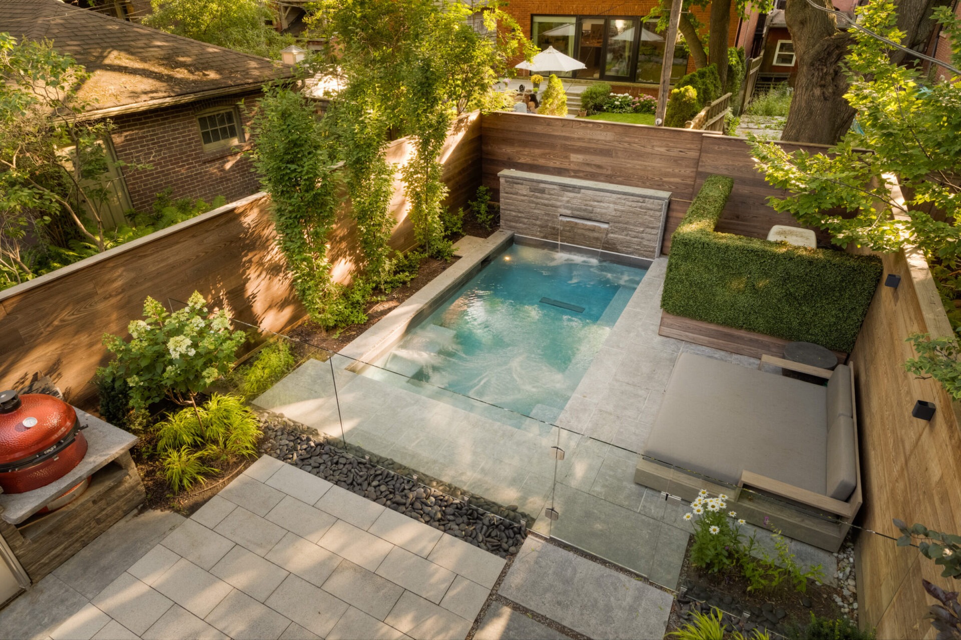 Cozy backyard with a small pool, lounge area, greenery, and a barbecue grill. A person is partially visible in the background.