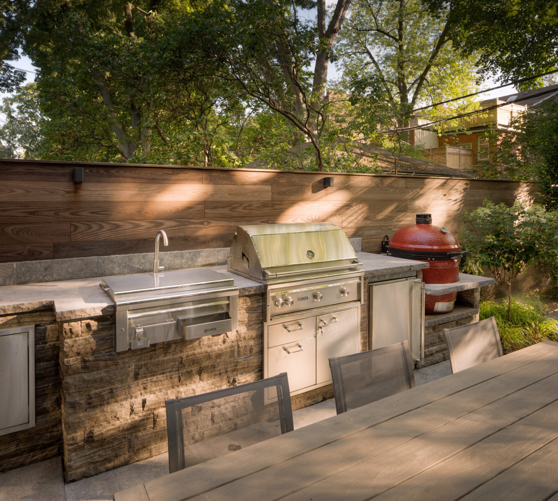 Outdoor kitchen with a sink, grill, and red ceramic cooker on a stone counter. Surrounded by trees and wooden fence.