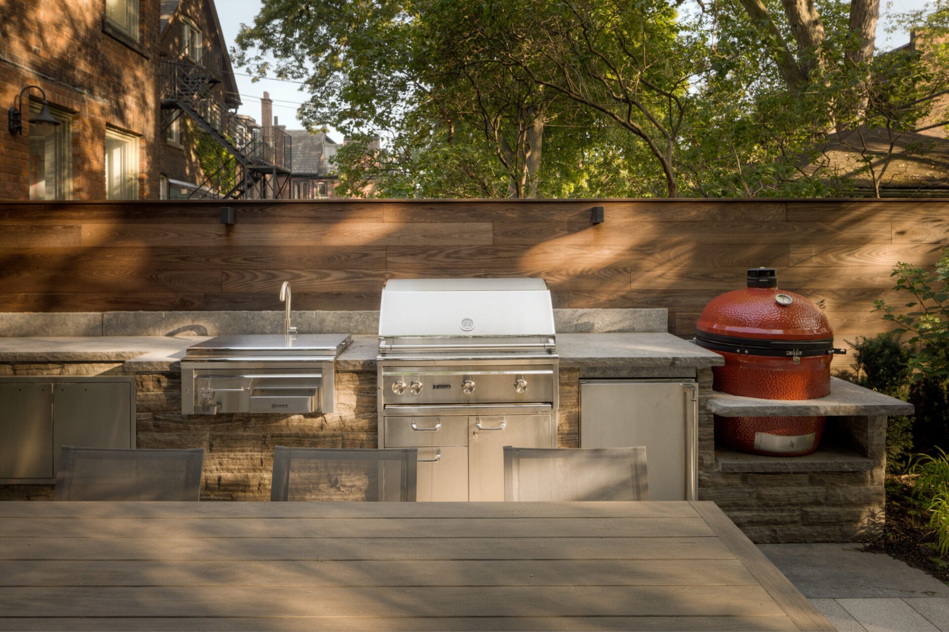 Outdoor kitchen with stainless steel appliances, wooden privacy fence, and a red grill. Surrounding area features trees and brick buildings.