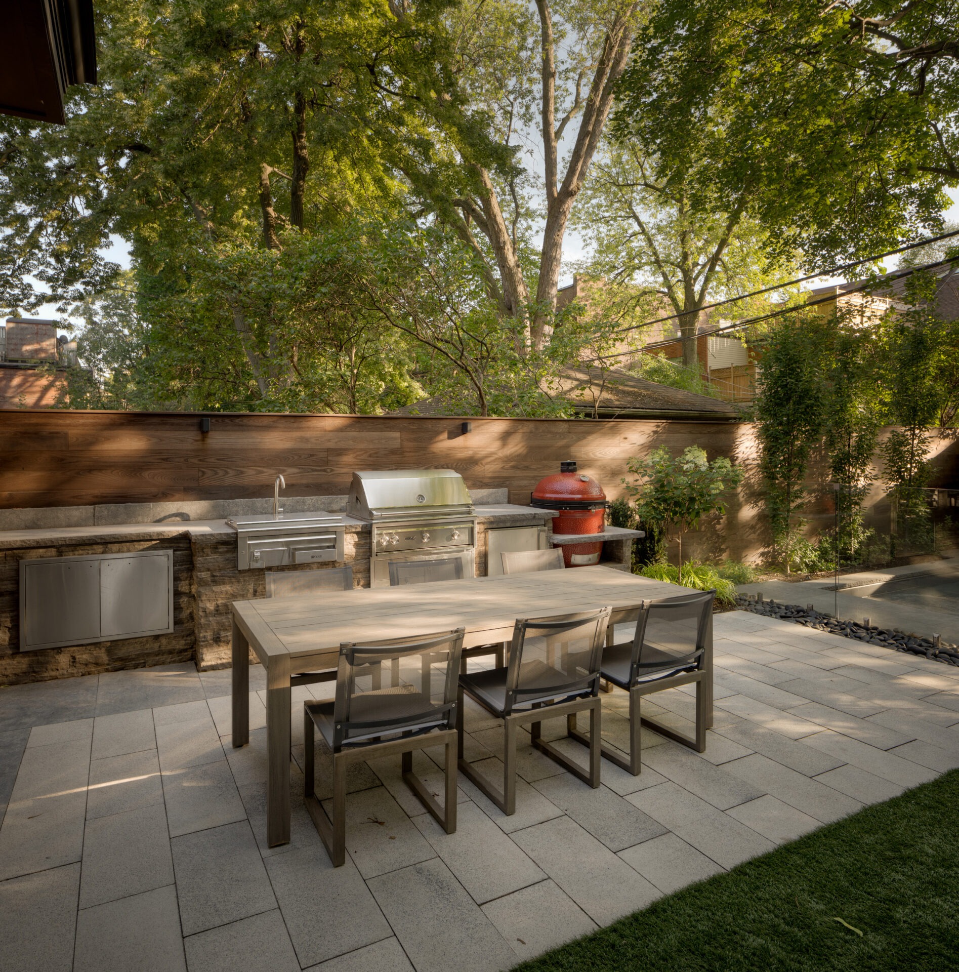 A modern outdoor kitchen with a grill, dining table, and chairs. Surrounded by trees and a wooden fence in a backyard setting.